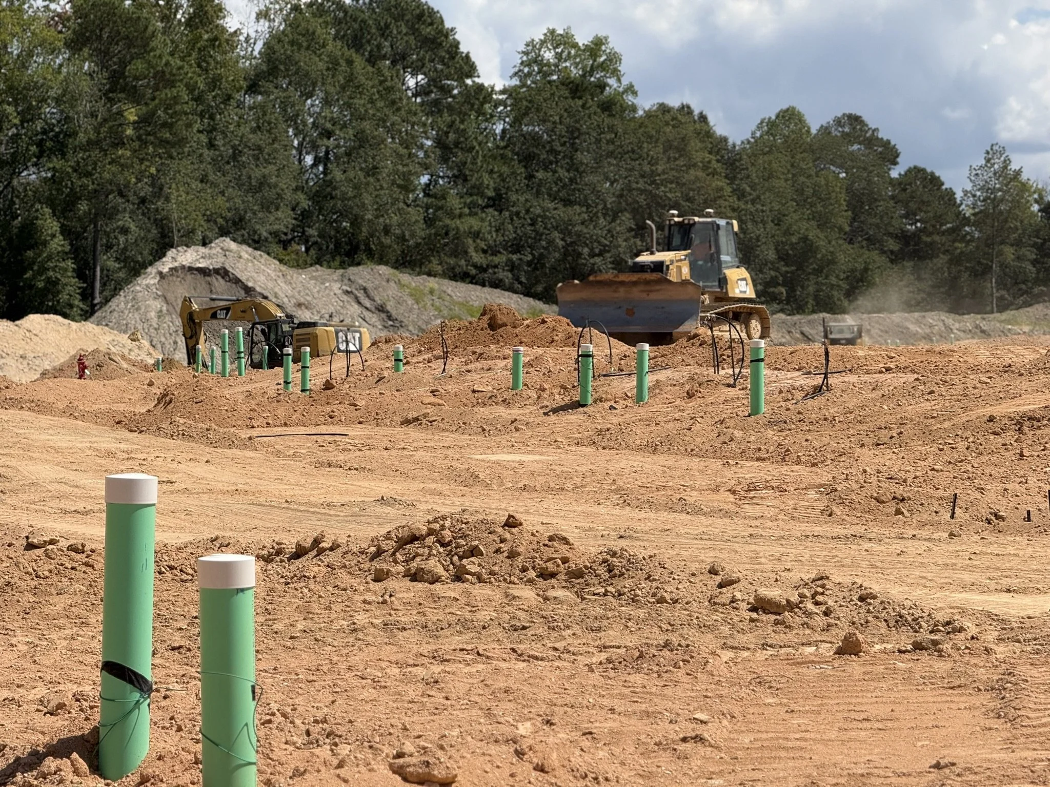 Construction site with bulldozer and mini excavator working on dirt, with green and white pipes installed in the ground, and trees in the background under partly cloudy sky.