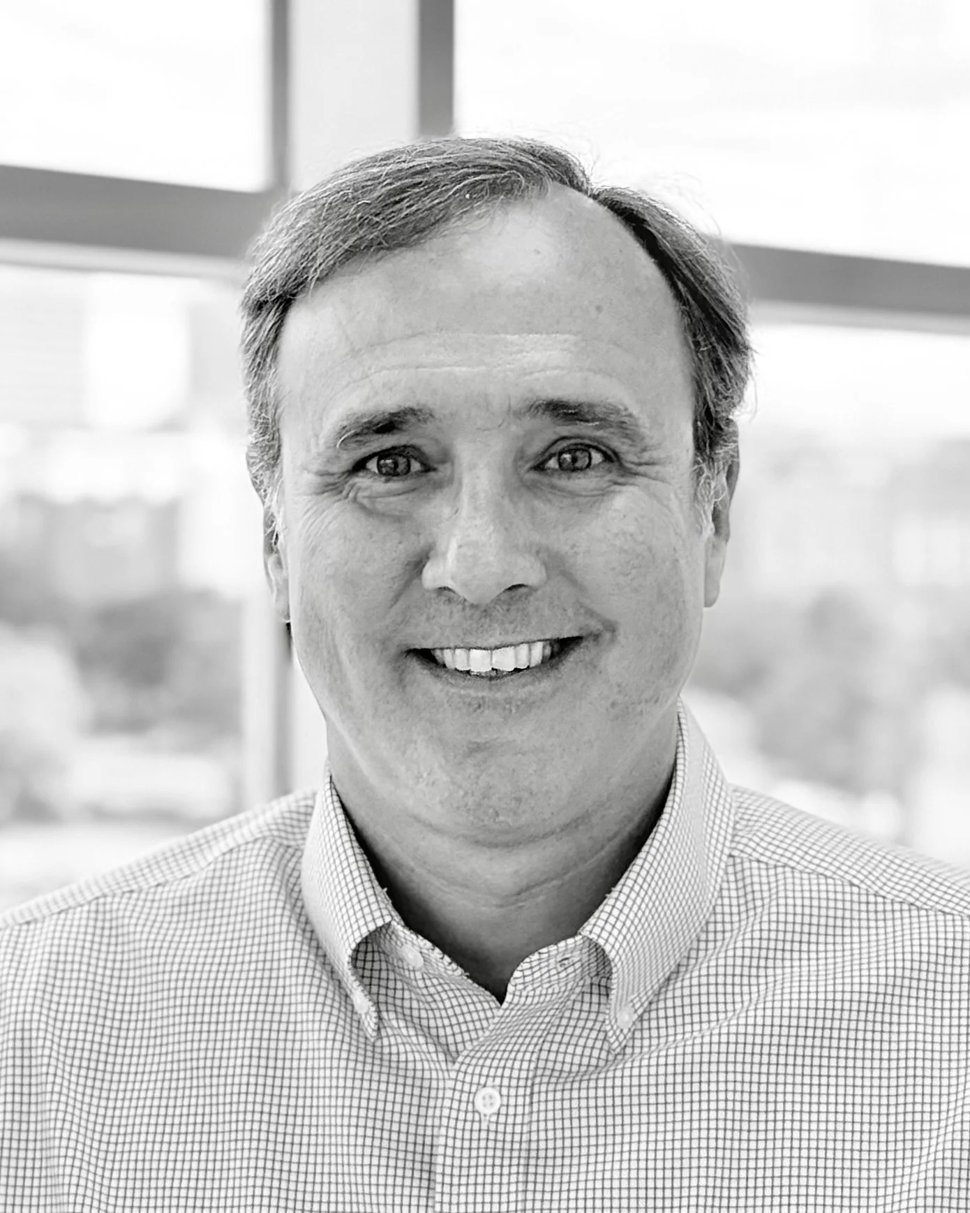 Black and white photo of a smiling man wearing a checkered collared shirt, with a blurred window background.