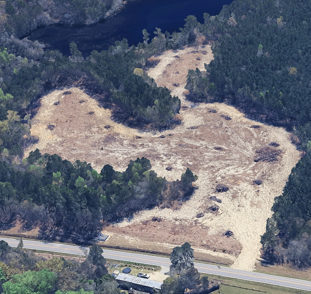 A cleared land area with winding dirt paths surrounded by dense trees, near a two-lane road at the bottom of the image.