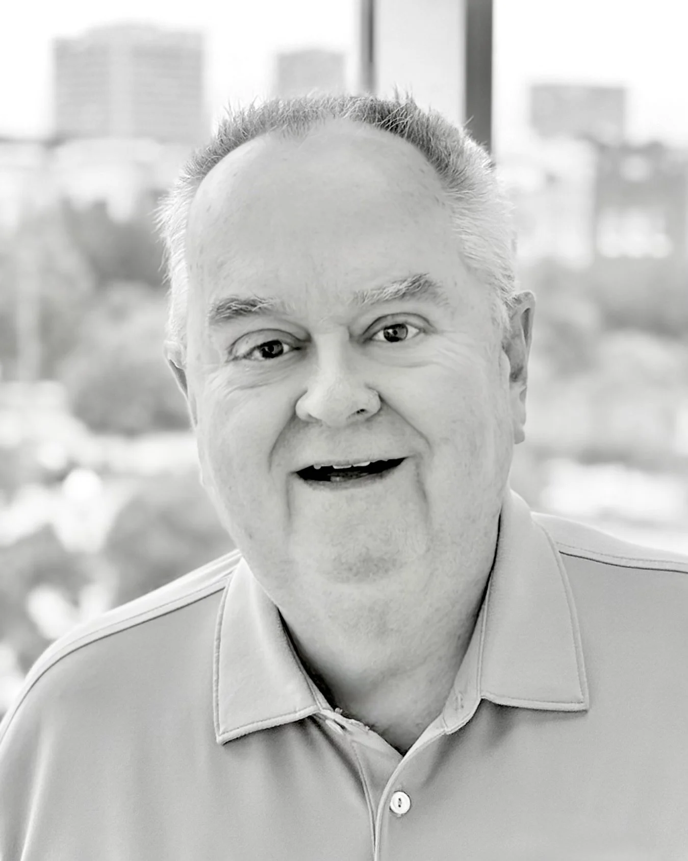 Black and white photo of a middle-aged man with short hair, smiling, wearing a collared shirt, with a cityscape window background.