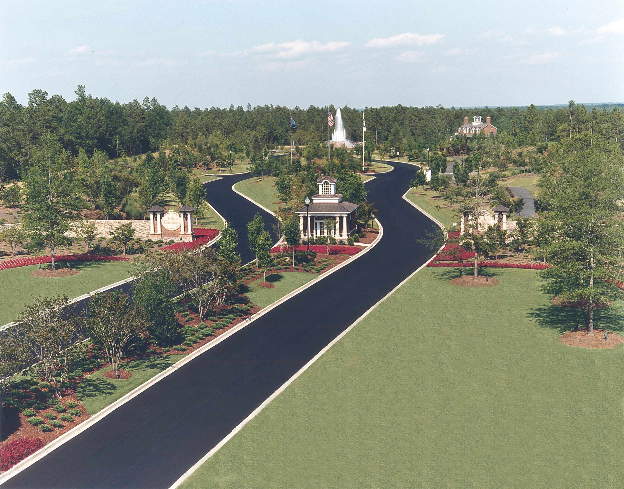 A winding black asphalt road with white borders leading into a park with green grass, trees, flower beds, a fountain, and a small building with columns in the center. Flags on poles are visible in the background, and a house is seen in the distance.