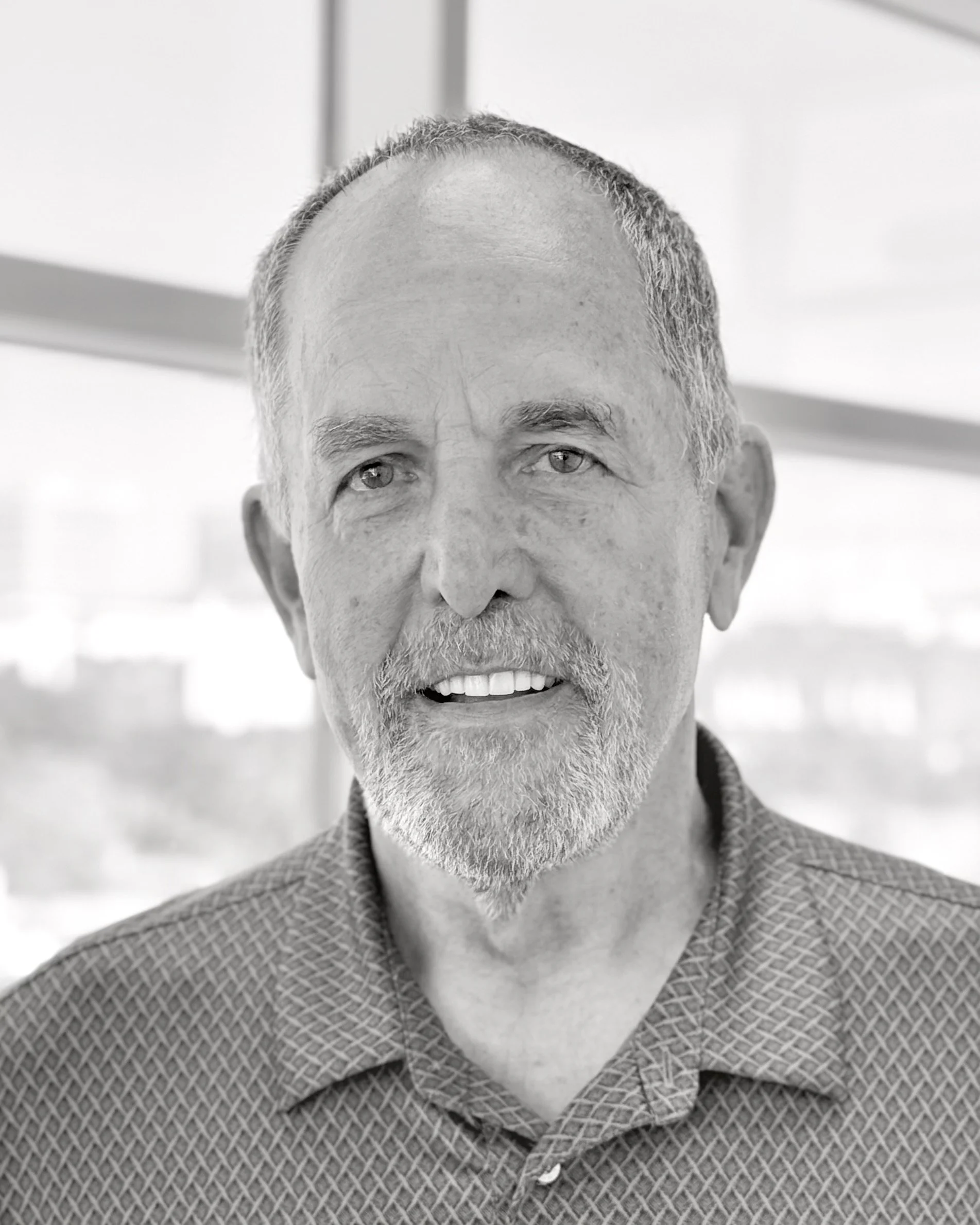 Black and white portrait of an older man with short gray hair, beard, and mustache, wearing a patterned collared shirt, standing indoors near a large window.