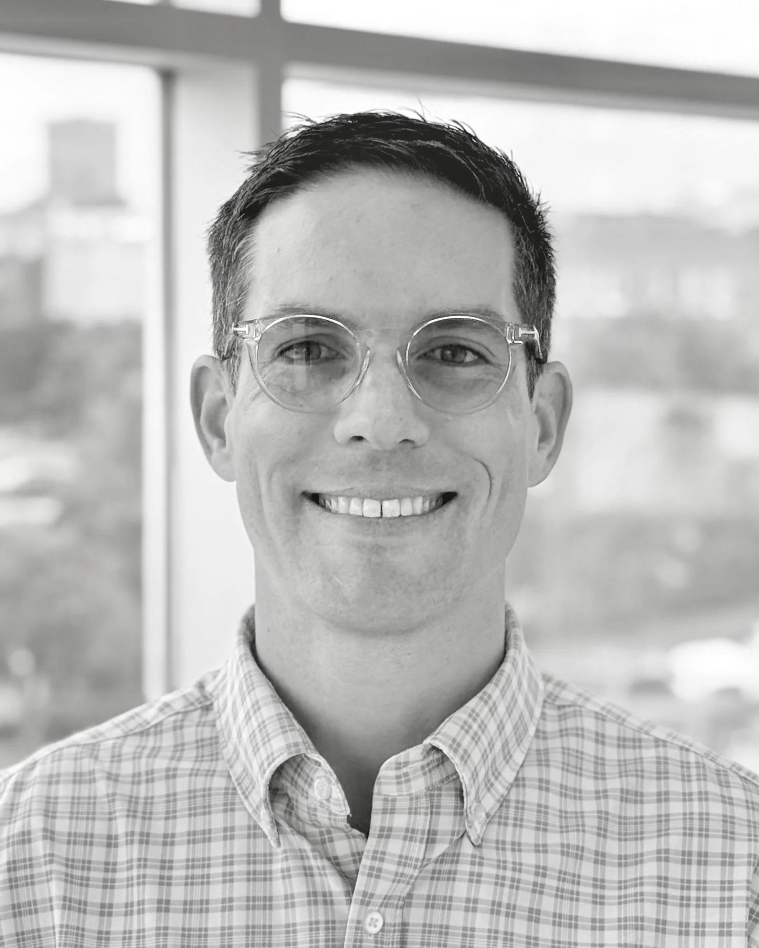 A smiling man with short dark hair, glasses, and wearing a checkered shirt, standing indoors near a window.
