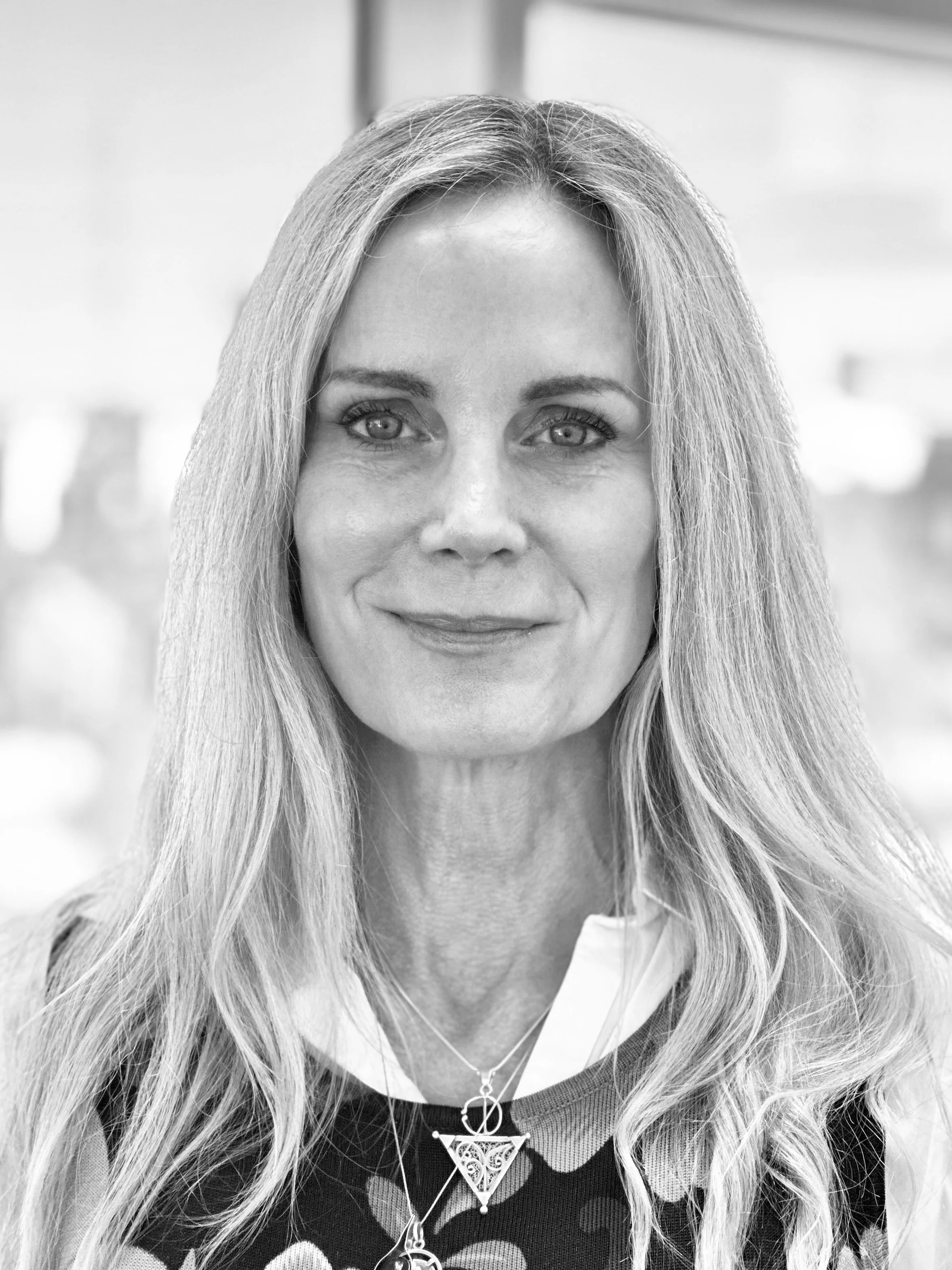 Black and white photo of a woman with long, wavy hair, smiling slightly, wearing layered necklaces and a patterned top.