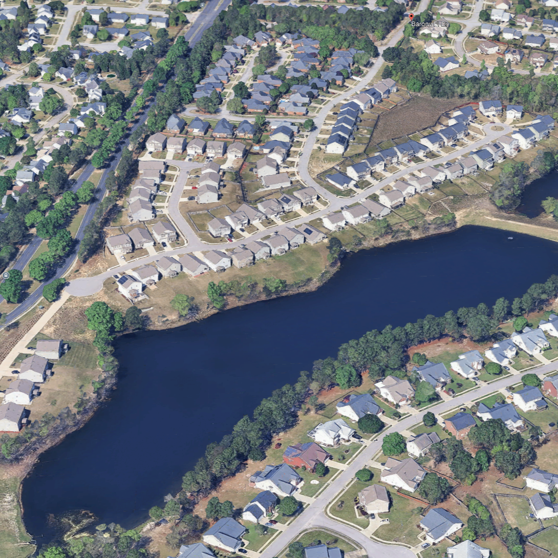 Aerial view of a suburban neighborhood with houses, roads, trees, a lake, and green spaces.