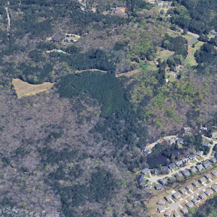 Aerial view of a wooded area with a mix of dense forest and cleared land, surrounded by residential neighborhoods and winding roads.