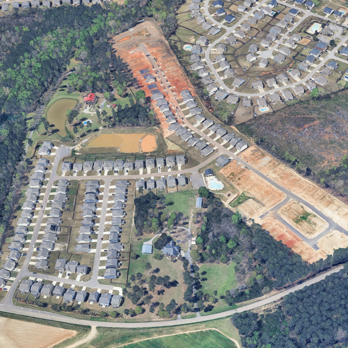 Aerial view of a suburban neighborhood with numerous houses, some with swimming pools, and undeveloped land with construction sites and wooded areas.