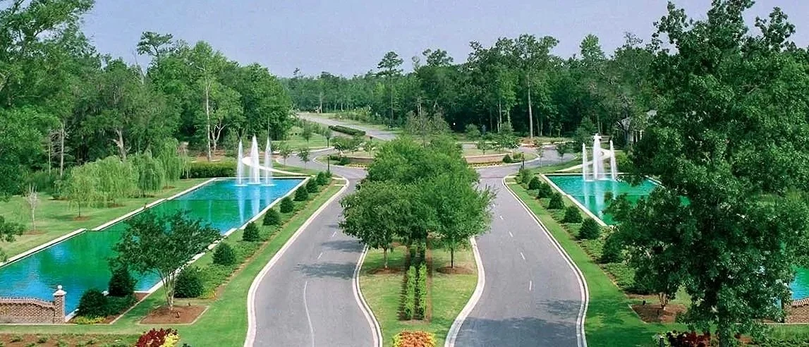 A landscaped driveway with fountains on either side and green trees surrounding a small park area in the middle.