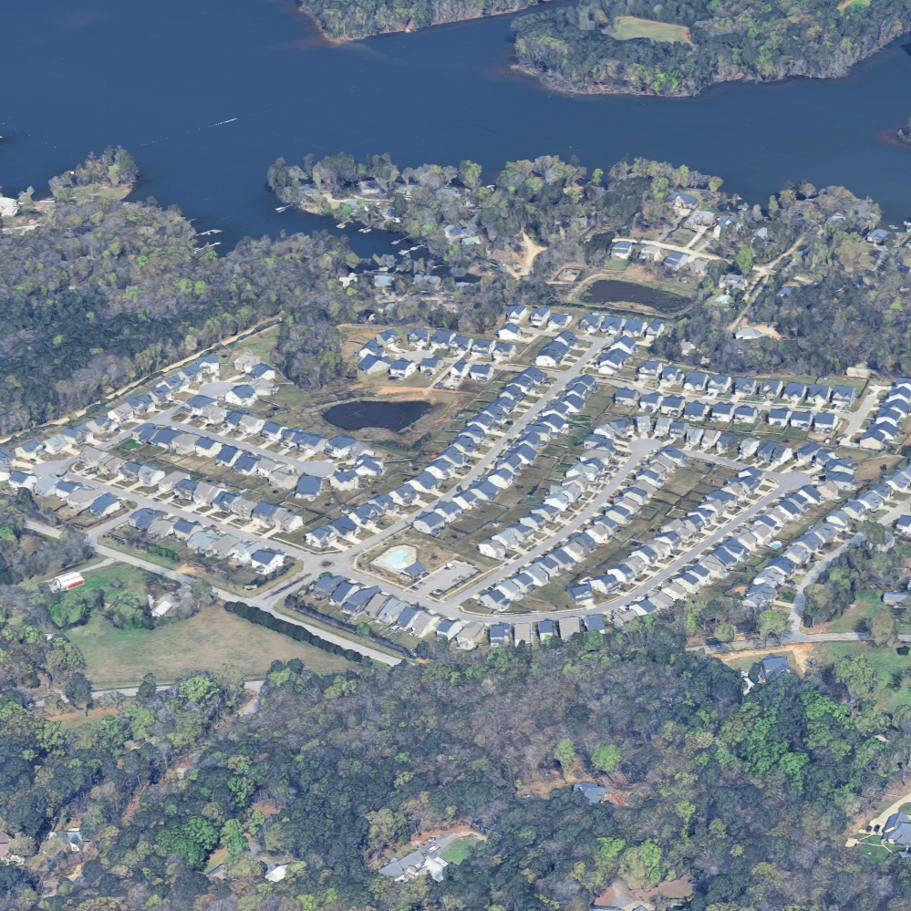 Aerial view of a neighborhood with houses, streets, ponds, and surrounding trees near a large body of water.
