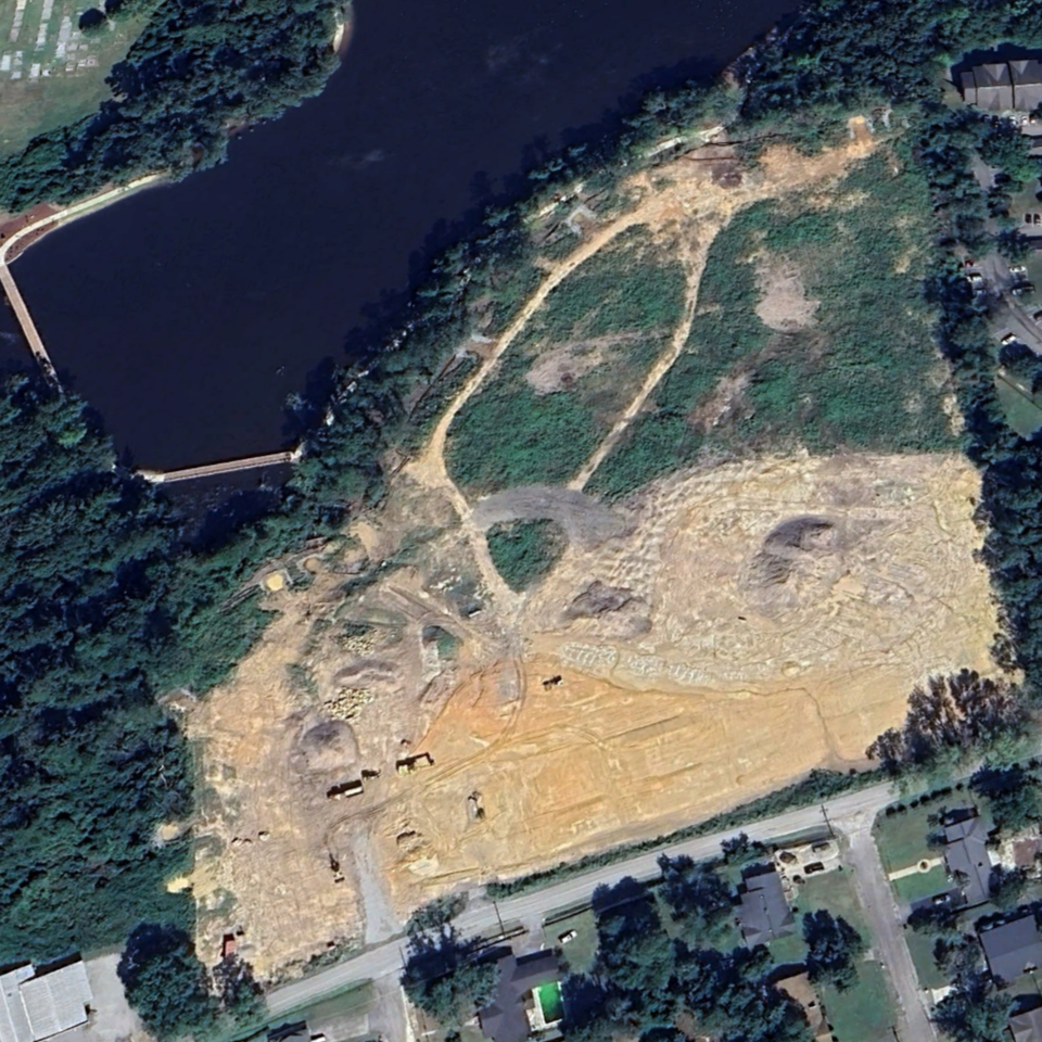 Aerial view of a construction site near a body of water with dirt, some green patches, and current excavation activity, surrounded by residential neighborhoods.