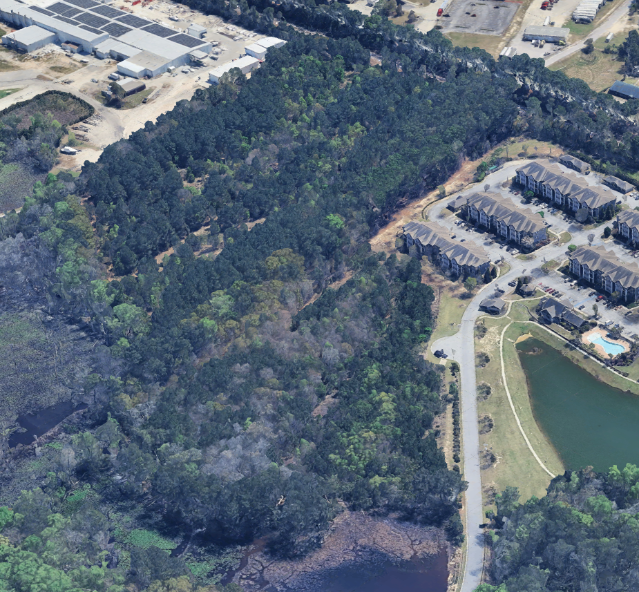 Aerial view of a wooded area with surrounding buildings, a pond, and an apartment complex with parking lot.