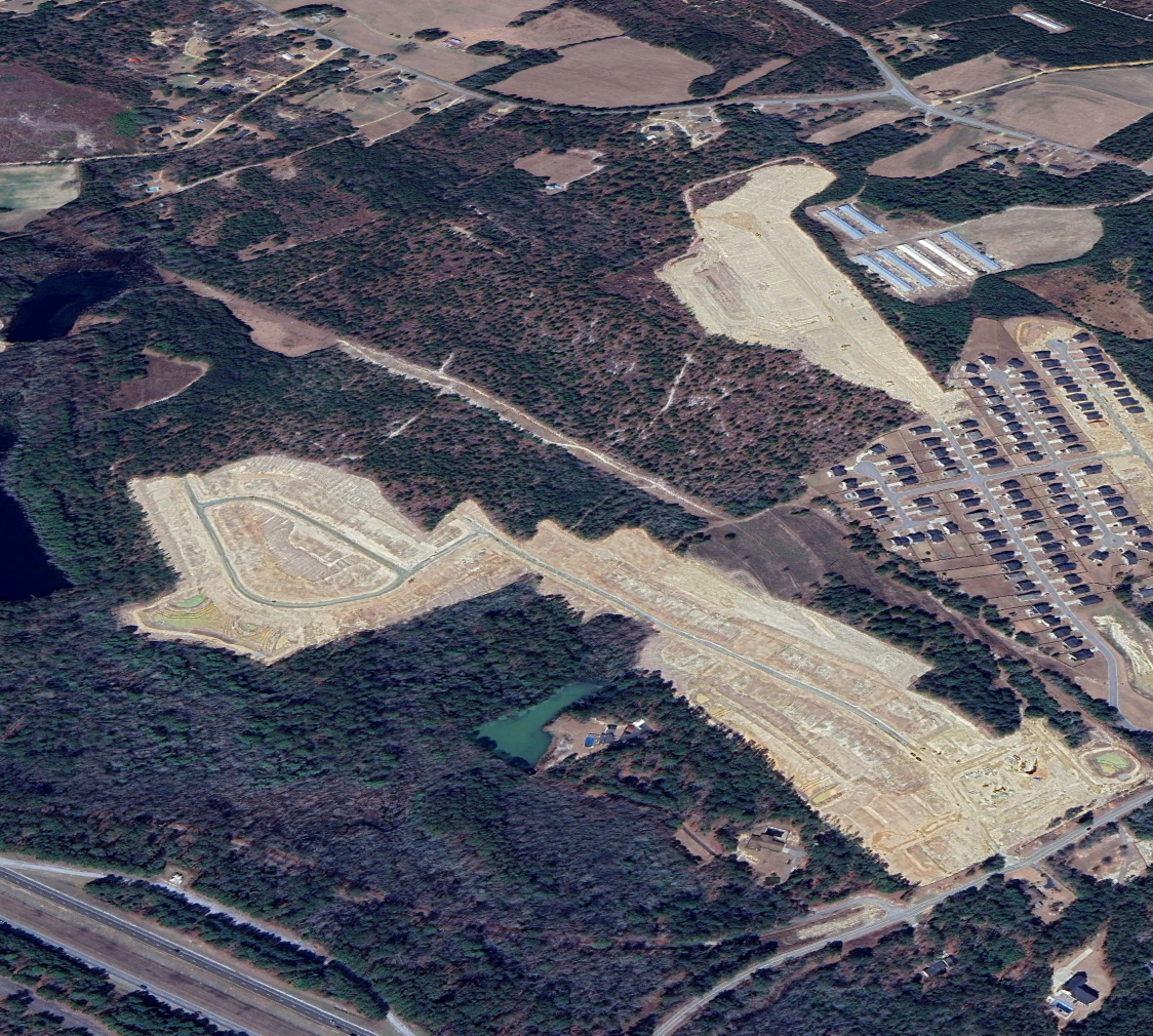 Aerial view of a construction site surrounded by wooded area, with roads, residential neighborhoods, and a small pond.