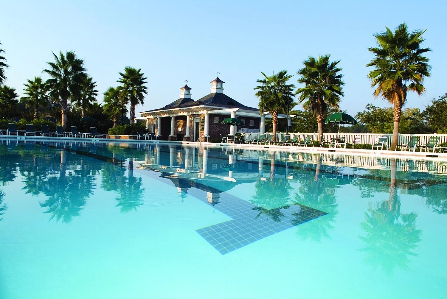 Empty outdoor swimming pool with clear blue water, surrounded by lounge chairs, umbrellas, palm trees, and a clubhouse building in the background under a clear sky.