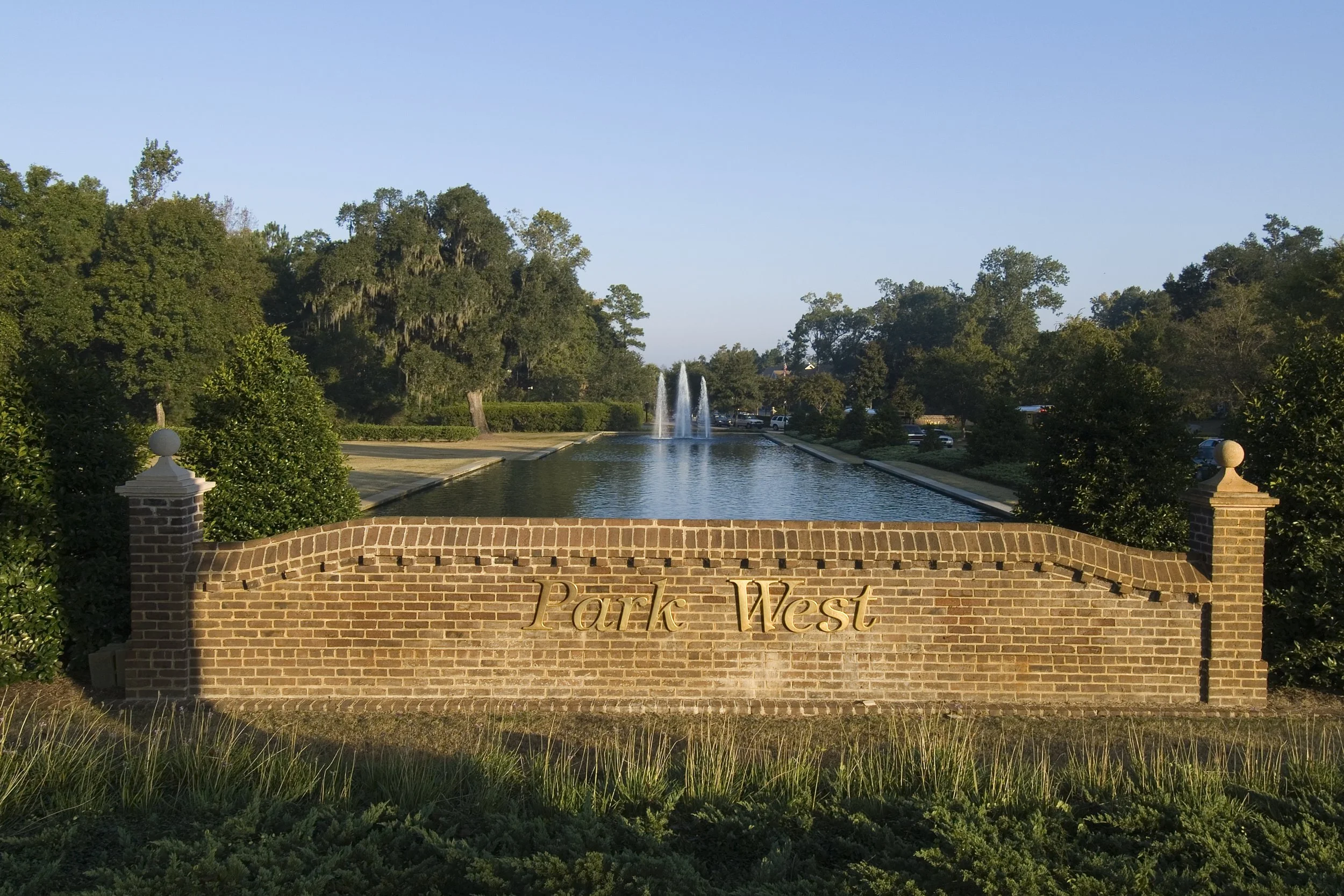 Brick entrance sign with gold lettering reading 'Park West' in front of a landscaped water feature and fountain, surrounded by trees.
