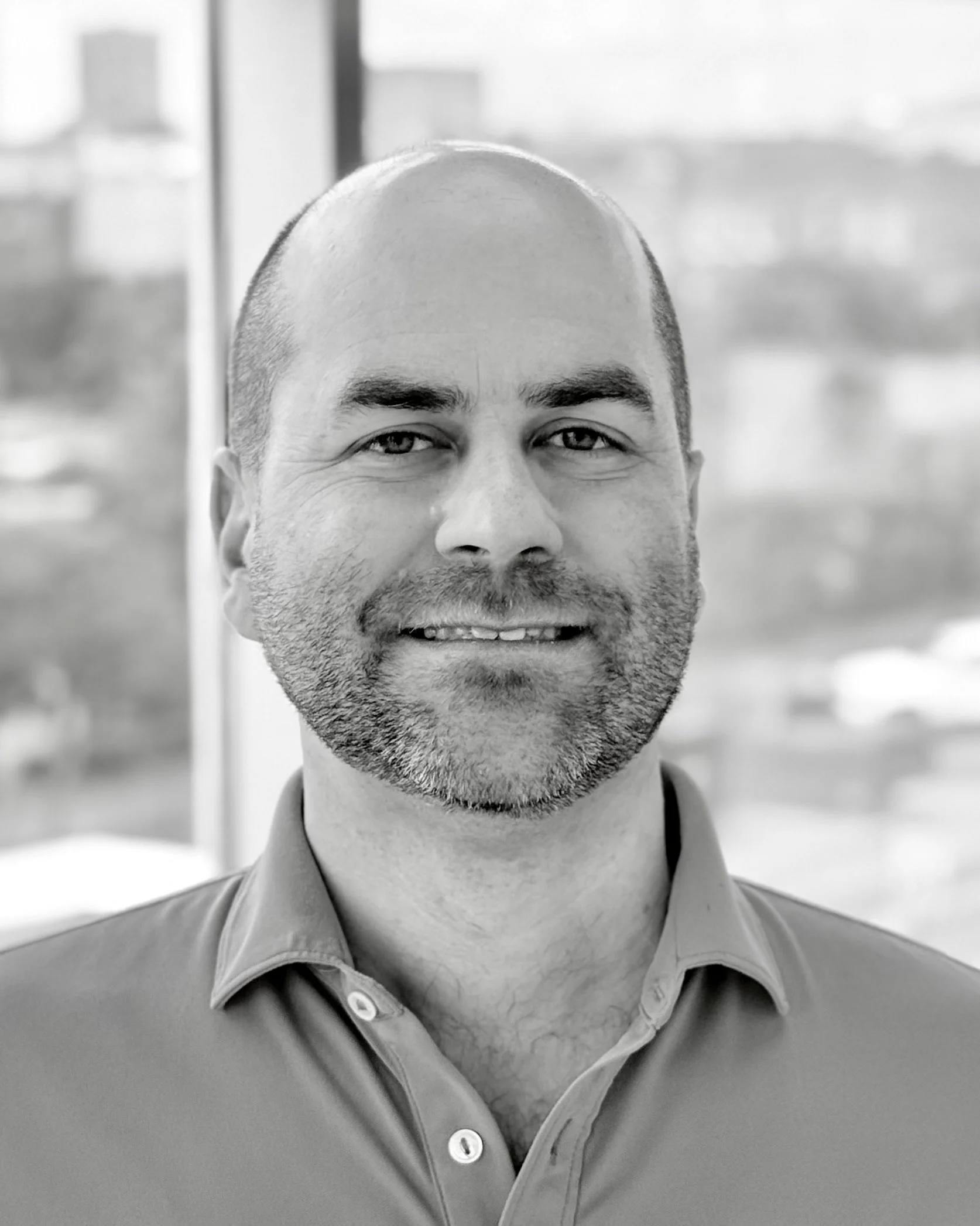 A smiling man with a beard, wearing a button-up shirt, in front of a window with blurred buildings outside.