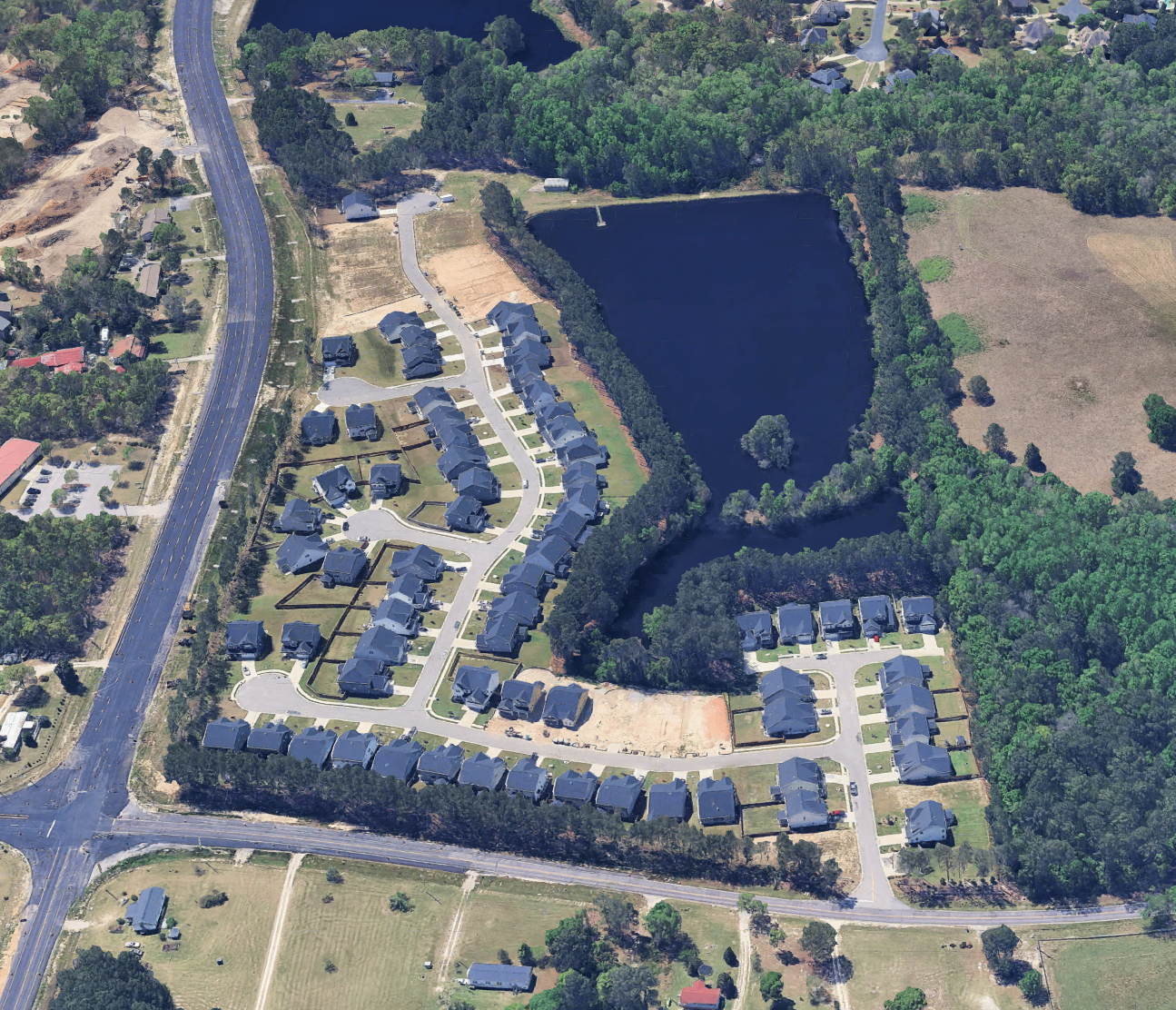 Aerial view of a residential neighborhood near a large pond surrounded by trees, with a curved street and several houses with dark roofs. A highway is visible on the left side of the image, and there are some open fields and wooded areas around.