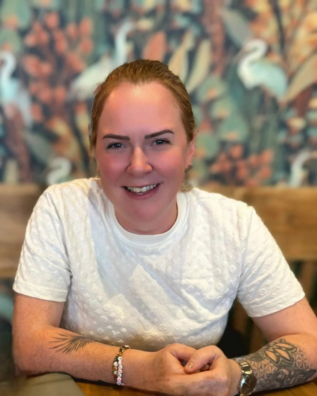 A woman with red hair and tattoos on her arms, wearing a white shirt, smiling at the camera in a restaurant setting with a patterned wall background.