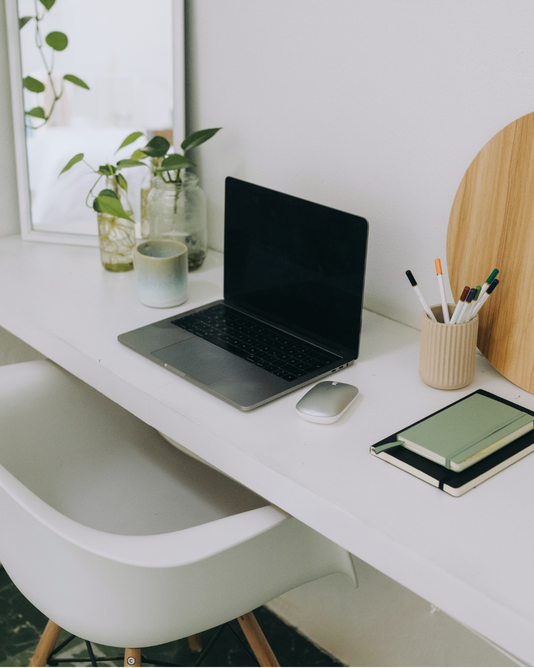 A clean, minimalist workspace with a white desk, an open laptop, a white mouse, notebooks, a beige pen holder with pens, potted plants, and a round mirror on the window sill.