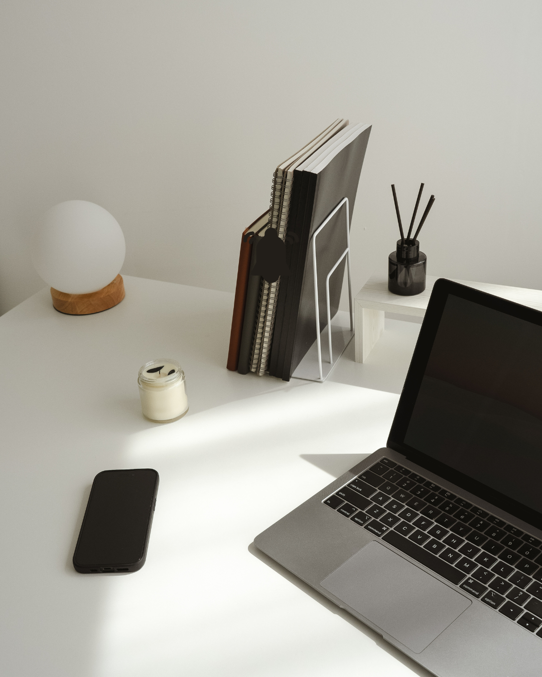 A minimalist workspace with a MacBook, smartphone, candle, a lamp, and storage racks with notebooks and folders, on a white desk against a plain light wall.