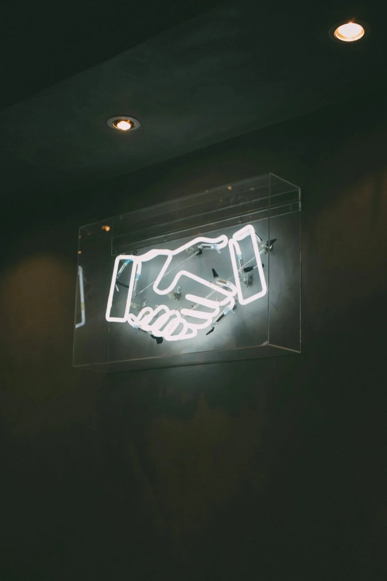 Neon sign in the shape of two shaking hands inside a clear enclosure, mounted on a dark wall with ceiling spotlights.