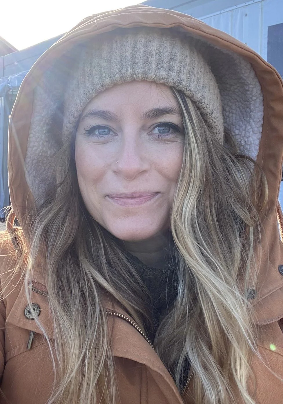 A woman with blue eyes and long wavy blonde hair wearing a beanie and a tan hooded jacket outdoors.