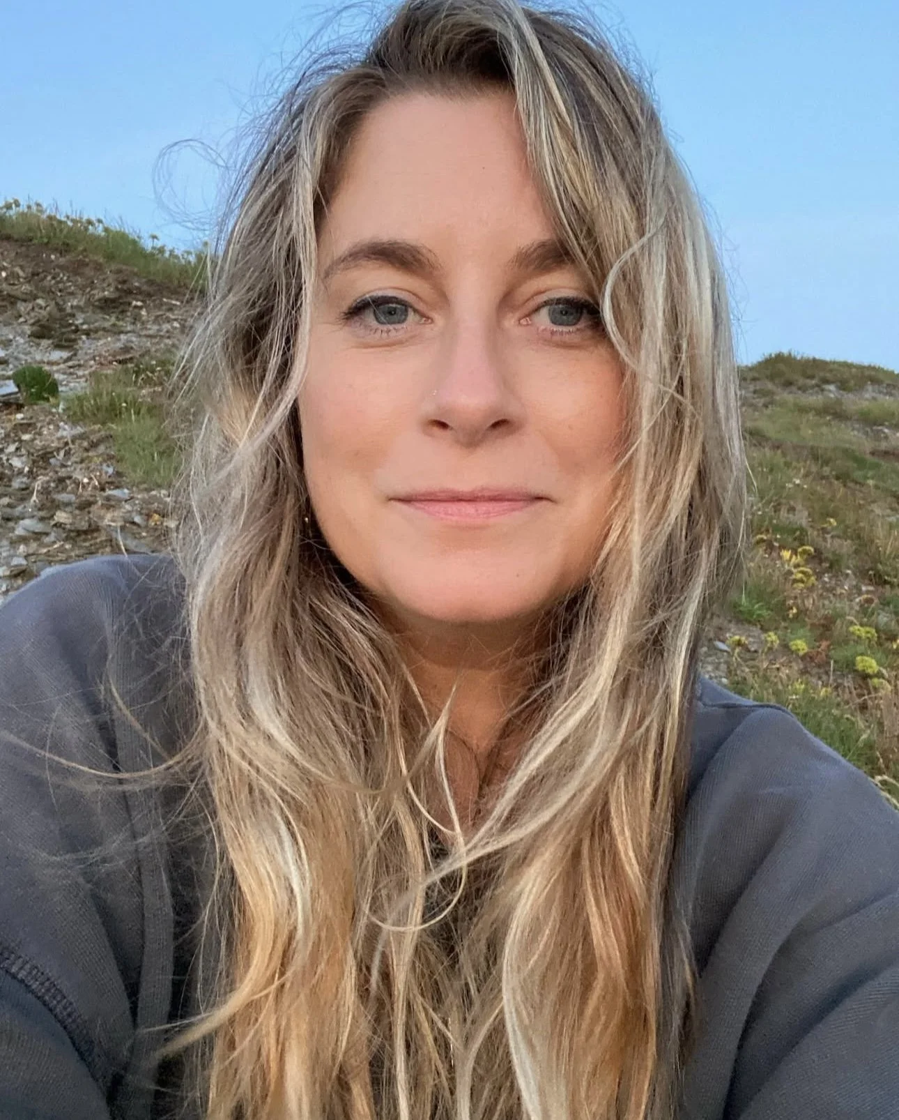 Close-up of a woman with long, wavy blonde hair and blue eyes, outdoors, with a grassy hillside and a clear blue sky in the background.