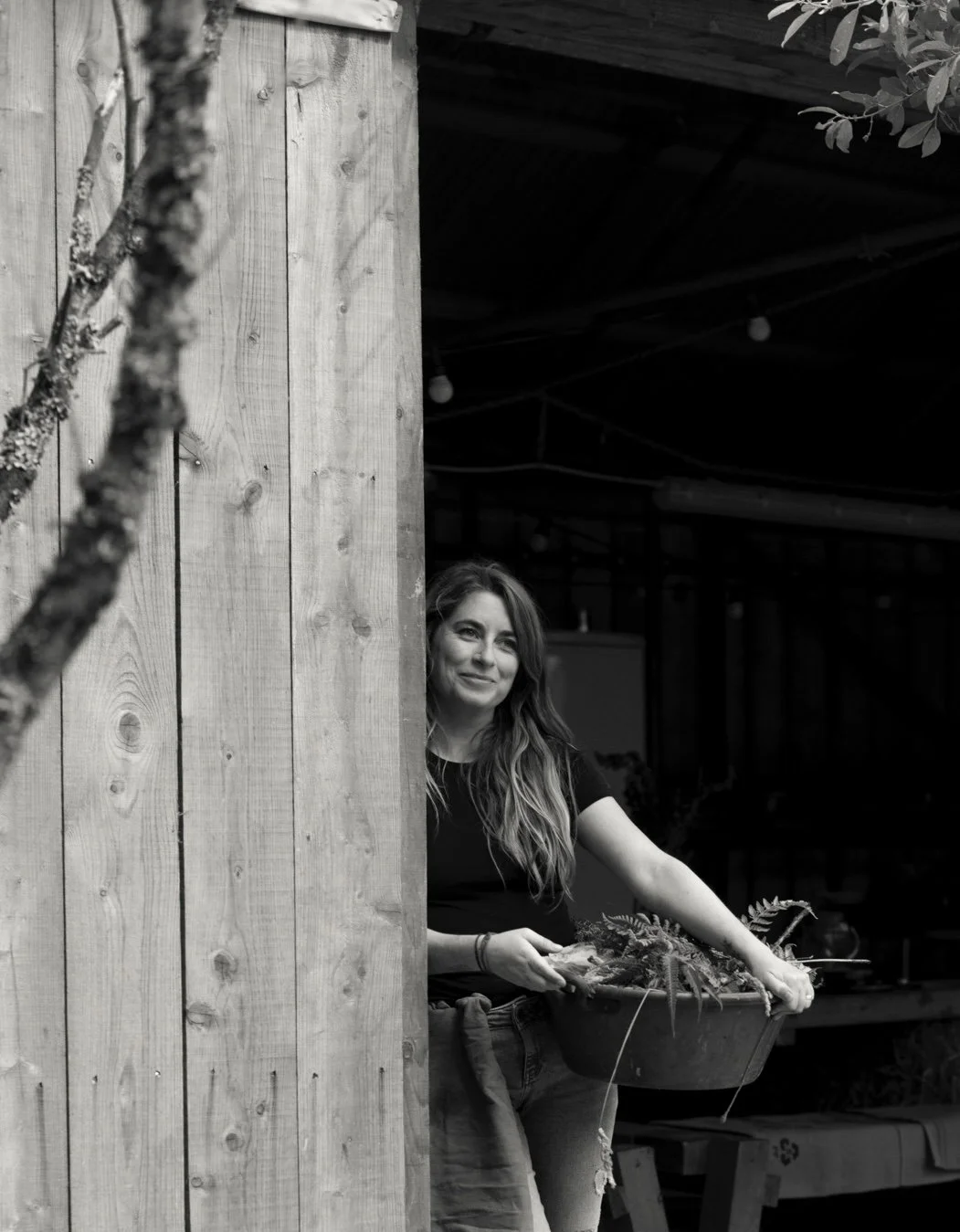 Woman with long hair holding a container of plants, standing next to a wooden wall, smiling