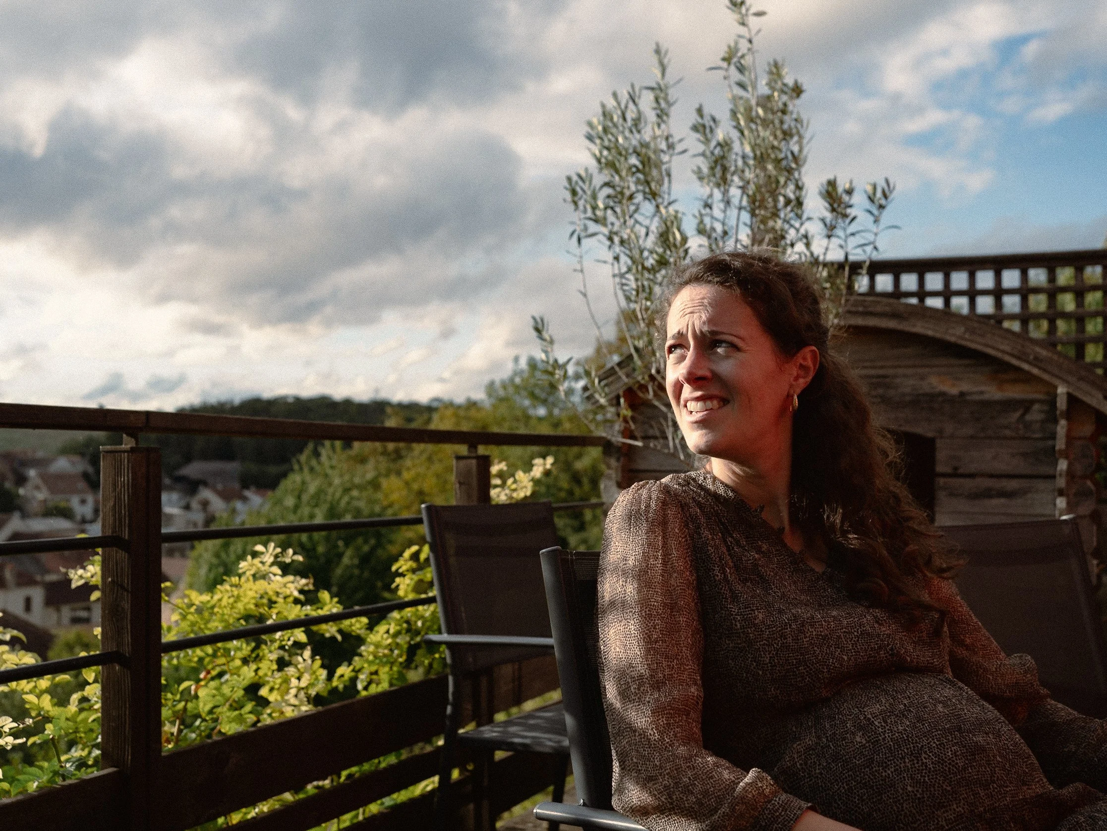 Une femme souriante assise sur une terrasse en bois avec un fond de ciel nuageux, des arbres et des maisons en arrière-plan, entourée de plantes et de fleurs à Guerville..