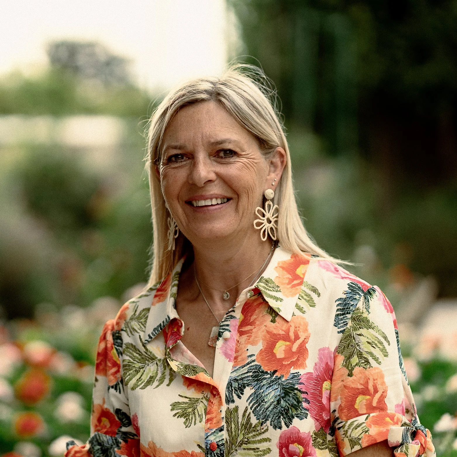 Une femme souriante portant une chemise à fleurs colorée, avec des boucles d'oreilles en forme de fleurs, en arrière-plan, le jardin et la maison de Claude Monet à Giverny.