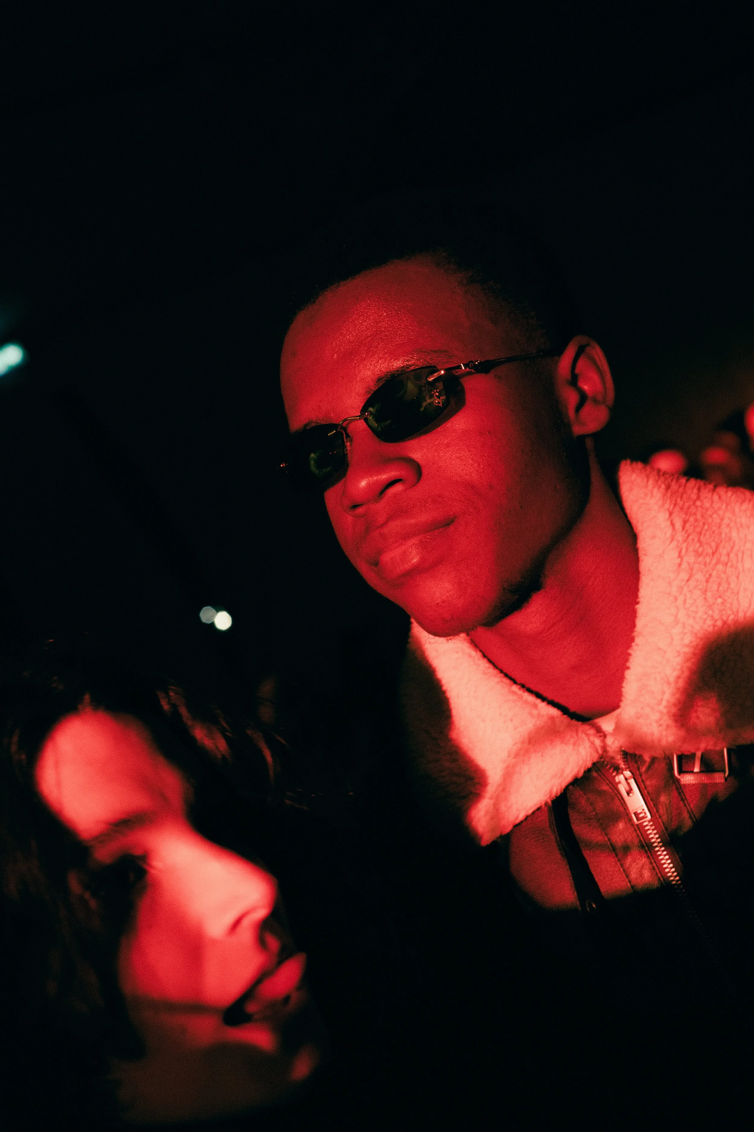 Une personne portant des lunettes de soleil et un manteau en peluche, sous un éclairage rouge, se trouvant dans un environnement sombre, avec une autre personne visible en contrebas.