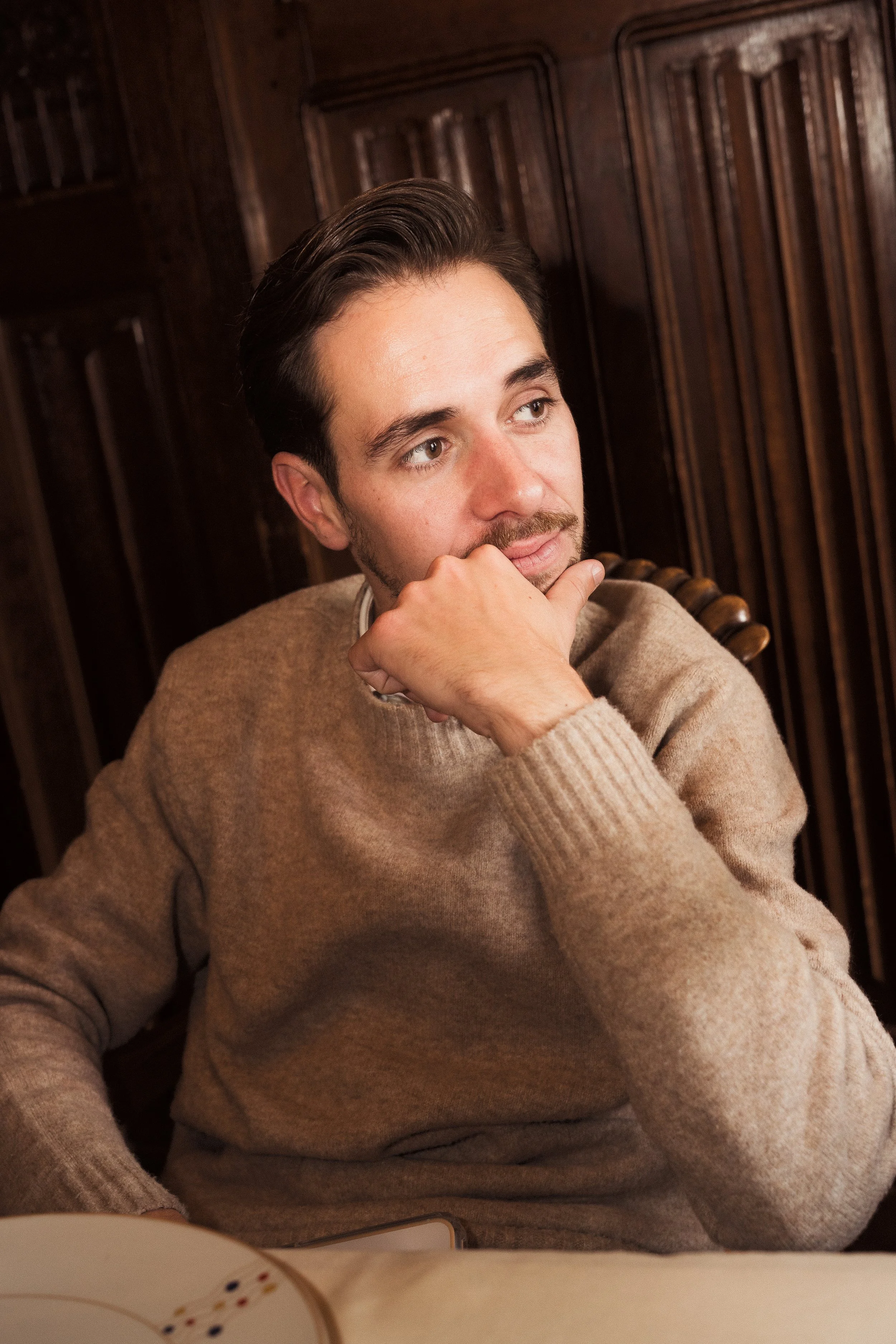 Un homme portant un pull beige, assis dans un intérieur en bois, semble pensif, avec la main sur le visage.