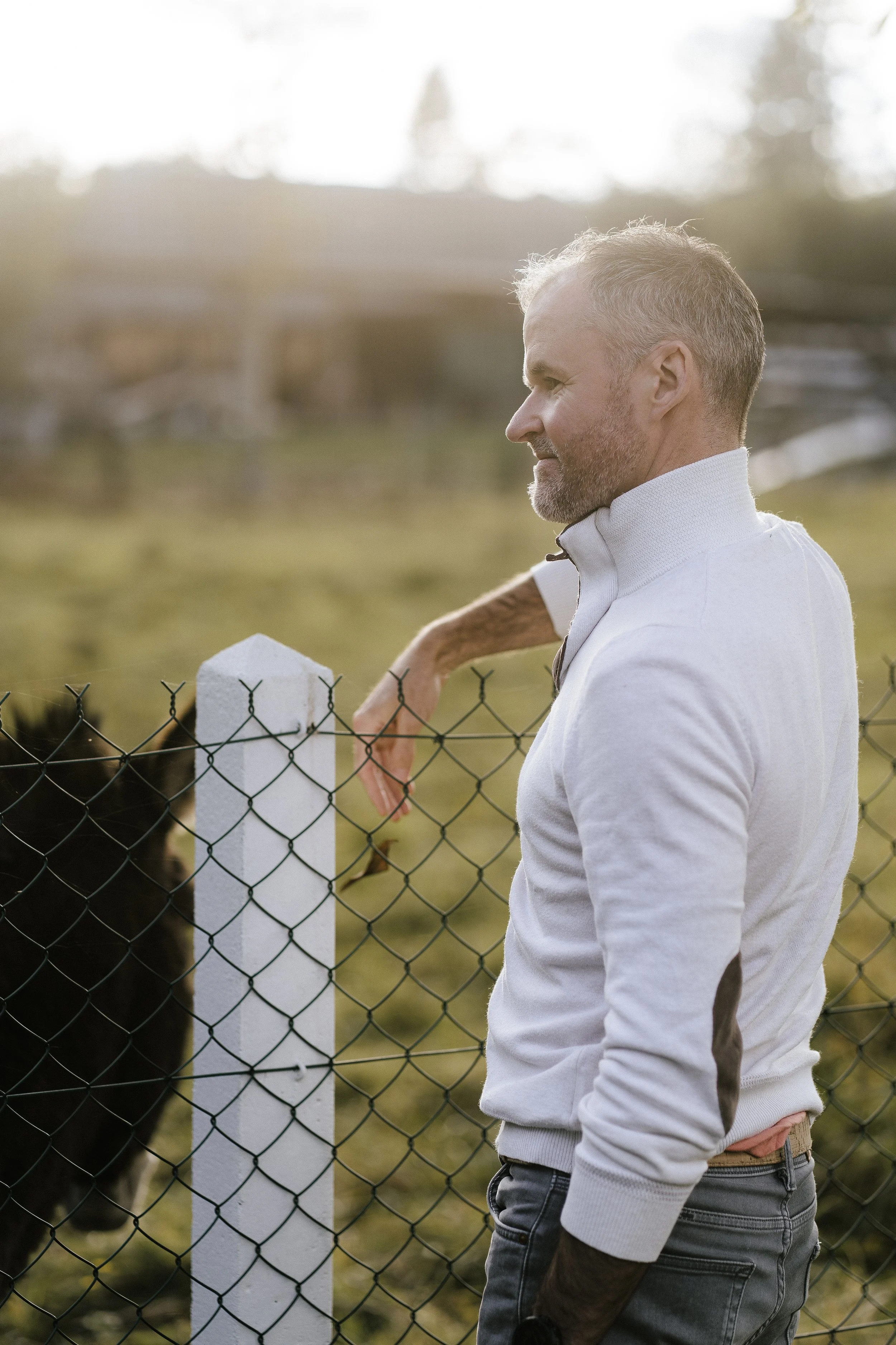 Un homme âgé, avec des cheveux gris, porte un pull blanc et regarde un animal derrière une clôture.