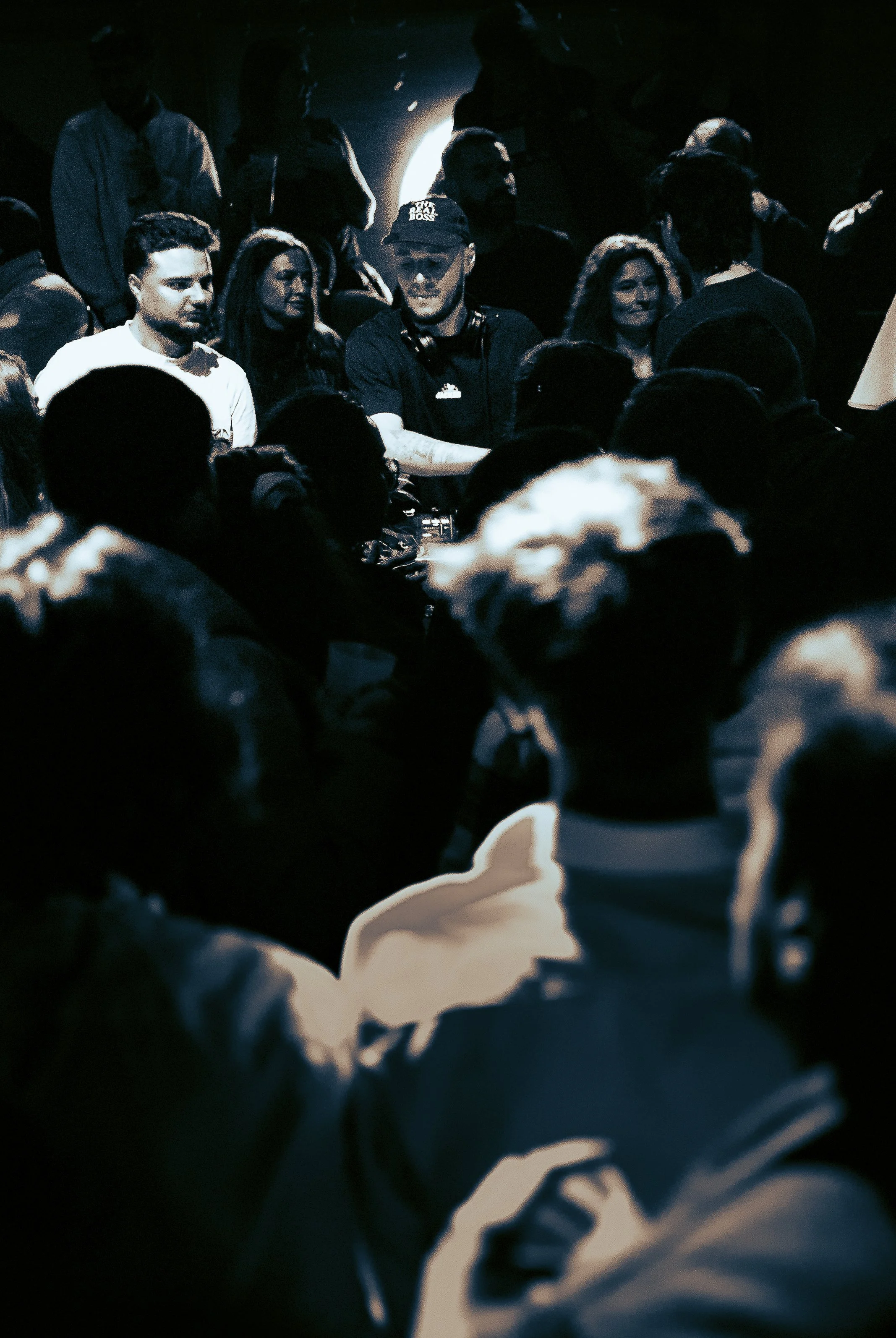 Un groupe de personnes réuni dans une pièce obscure, probablement lors d'une session de musique ou d'un événement social, avec un DJ en avant-plan portant une casquette noire.