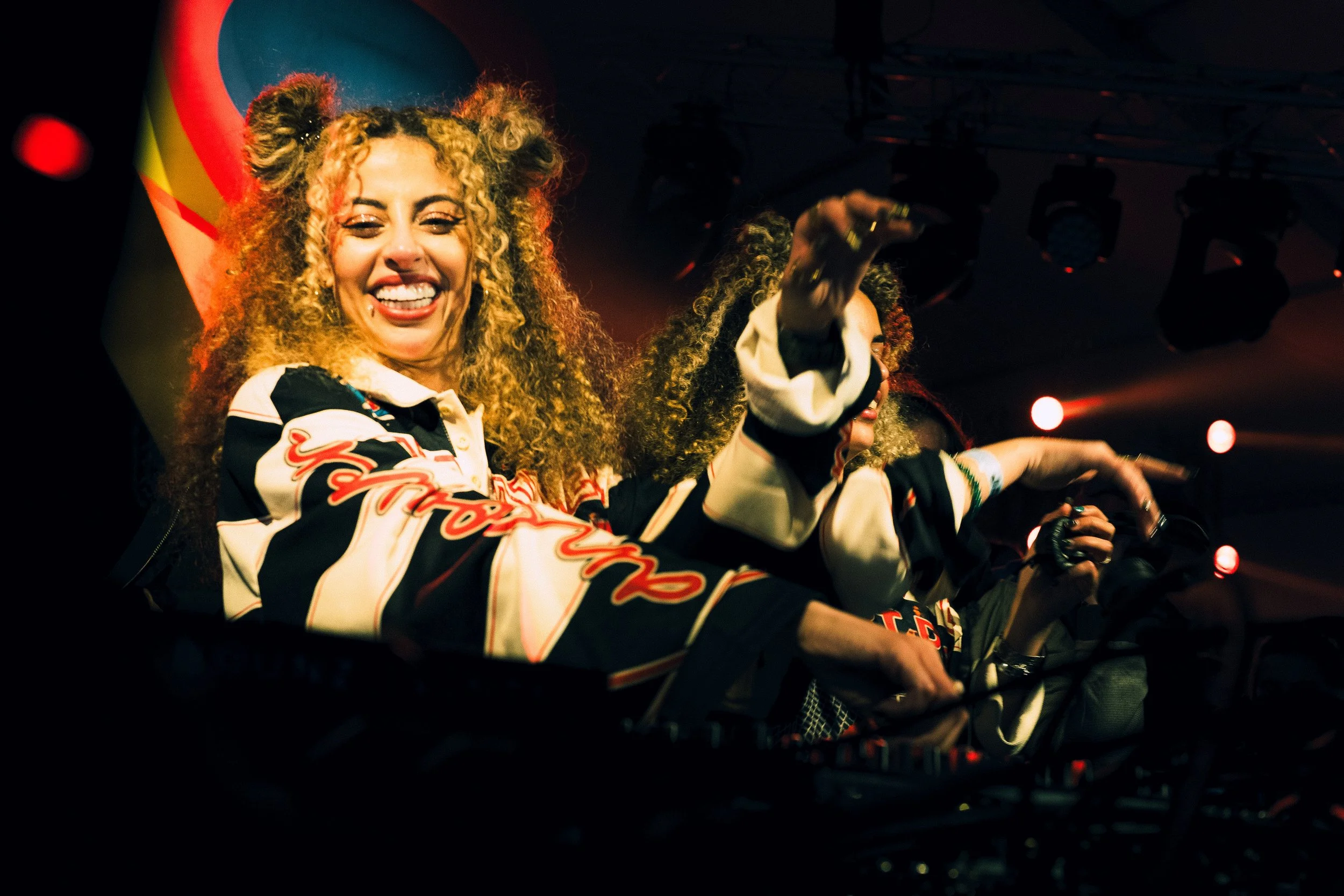 Deux femmes avec des cheveux bouclés et des tresses faisant la fête, souriantes, sur scène en illustration. Elle portent des vêtements colorés et sont entourées de lumières de scène. Pendant le festival Jacques Fromages.