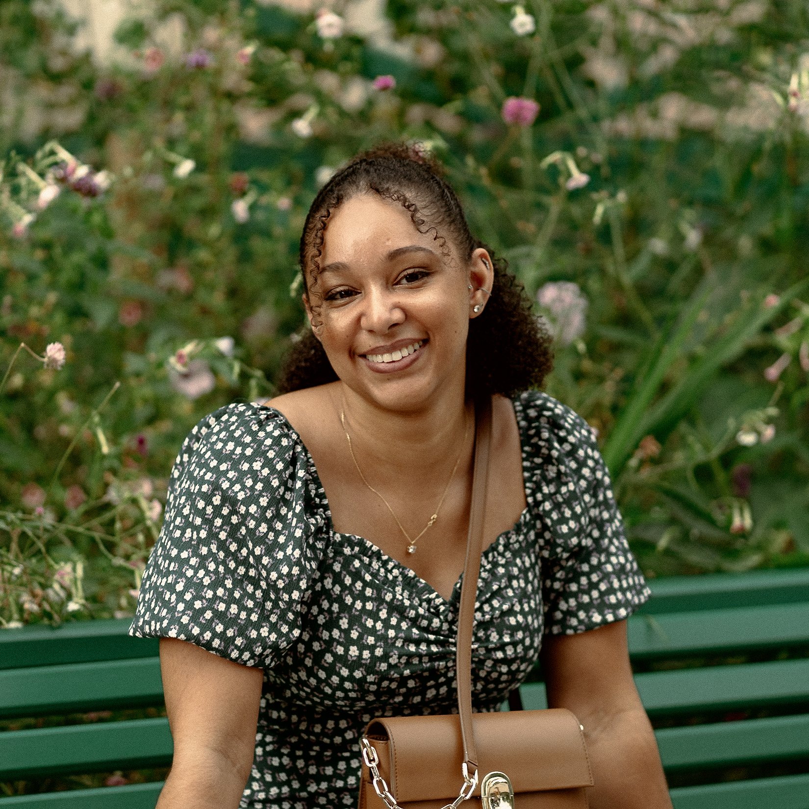 Une jeune femme souriante assise sur un banc vert dans un jardin fleuri, portant une robe à motifs et un collier, avec un sac beige posé sur ses genoux. En arrière-plan, le jardin et la maison de Claude Monet à Giverny.