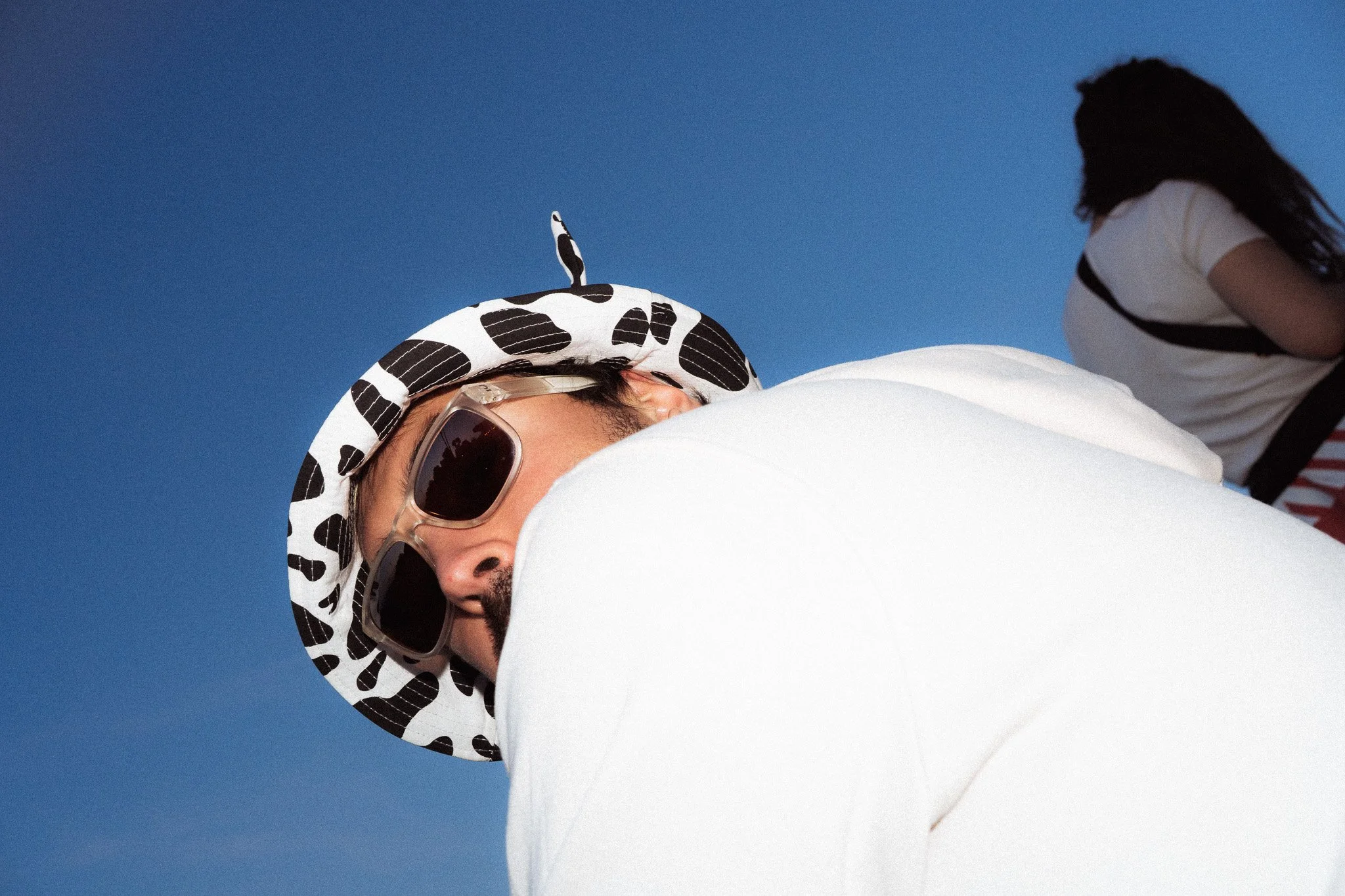 Une personne portant une casquette à motif noir et blanc, des lunettes de soleil, un t-shirt blanc et un pantalon blanc, vue de profil contre un ciel bleu, en position allongée au festival Jacques Fromages.