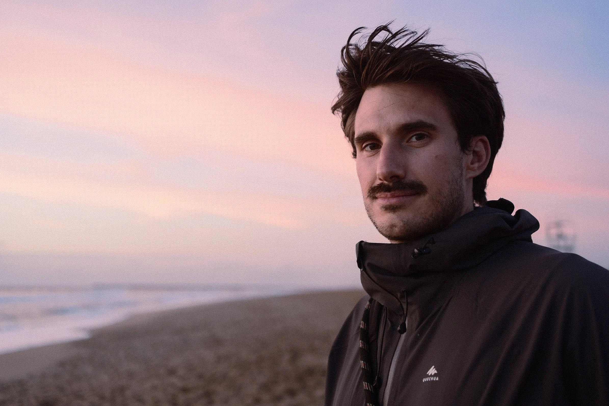 Un homme avec des cheveux bruns, barbe et moustache, portant une veste noire, regarde vers la caméra avec un ciel pastel au coucher du soleil en arrière-plan sur la plage d'Anglet au Pays Basque.