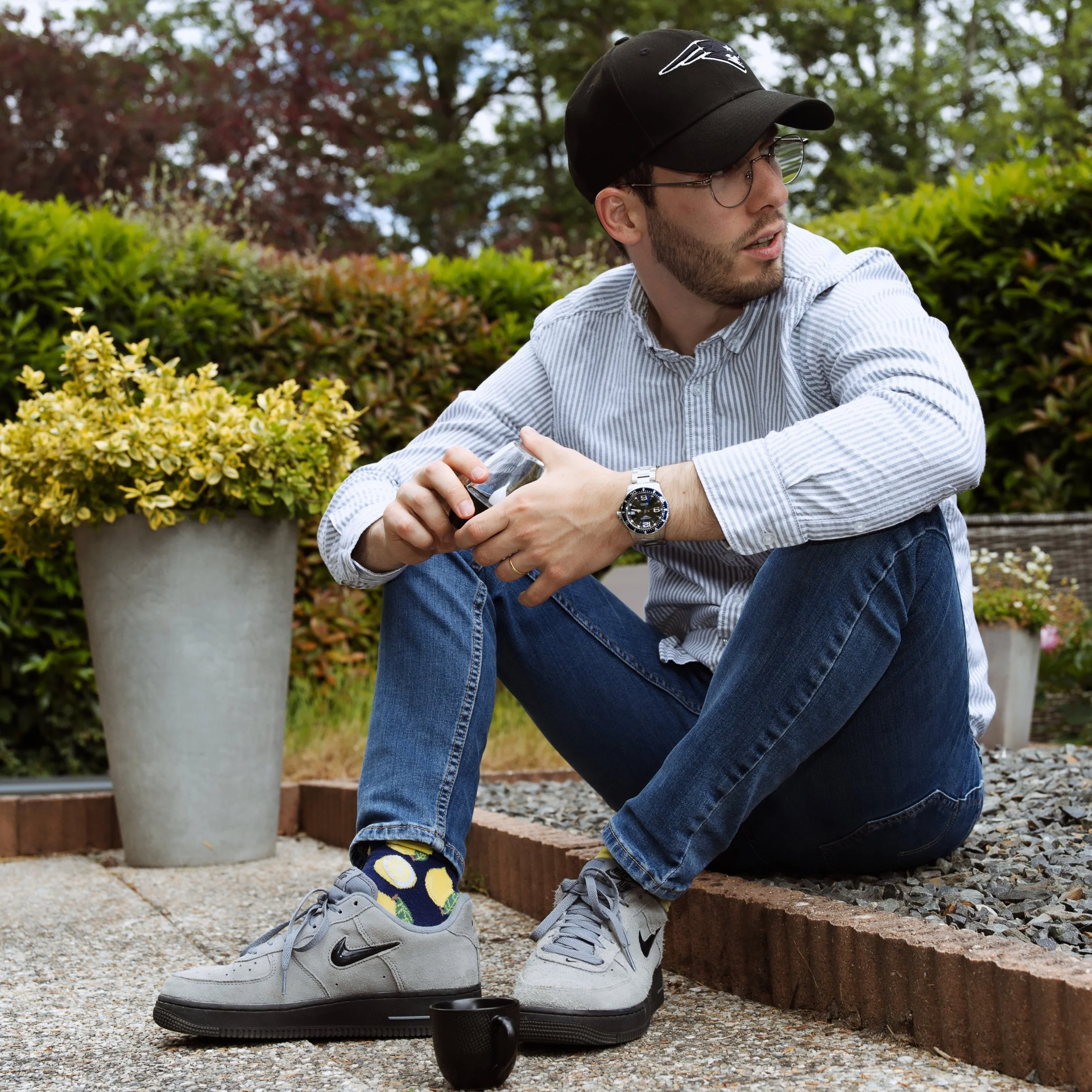 Un jeune homme assis par terre dans un jardin, portant une casquette noire, des lunettes, une chemise en denim, des jeans et des baskets Nike, avec un mug noir à côté, entouré de plantes et de buissons.