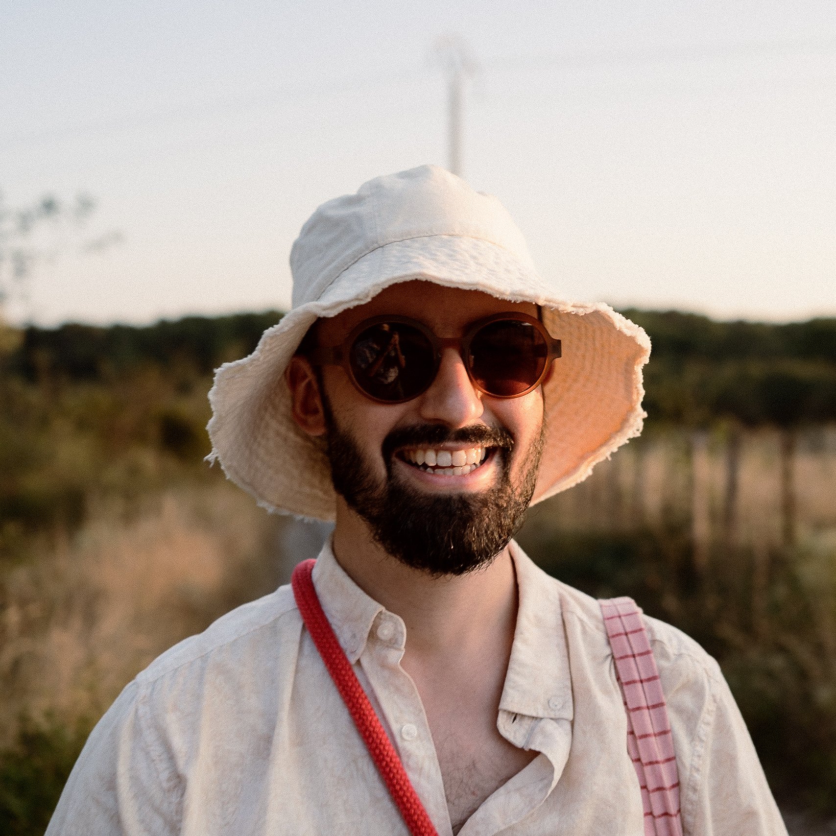 Un homme souriant porte un chapeau blanc, des lunettes de soleil rondes et une chemise blanche, avec un sac rose à ses épaules, dans un paysage naturel en plein air.