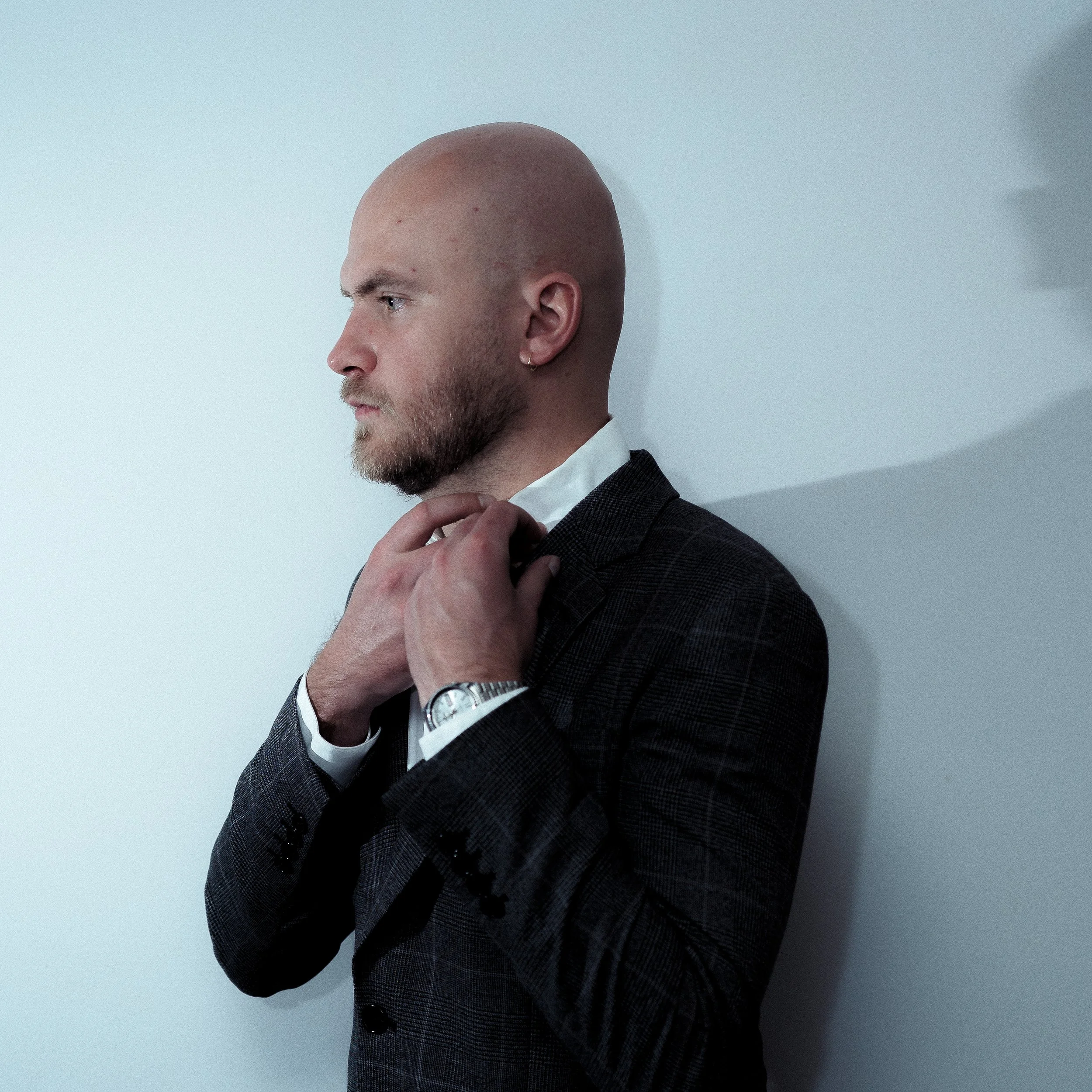 Homme coiffé d'une tête rasée, ajustant sa cravate, portant un costume sombre, avec un mur blanc en arrière-plan. Portrait Corportate.