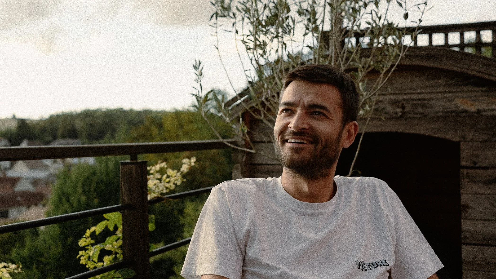 Un homme souriant assis sur une terrasse en bois avec un arrière-plan de nature et un ciel nuageux à Guerville.