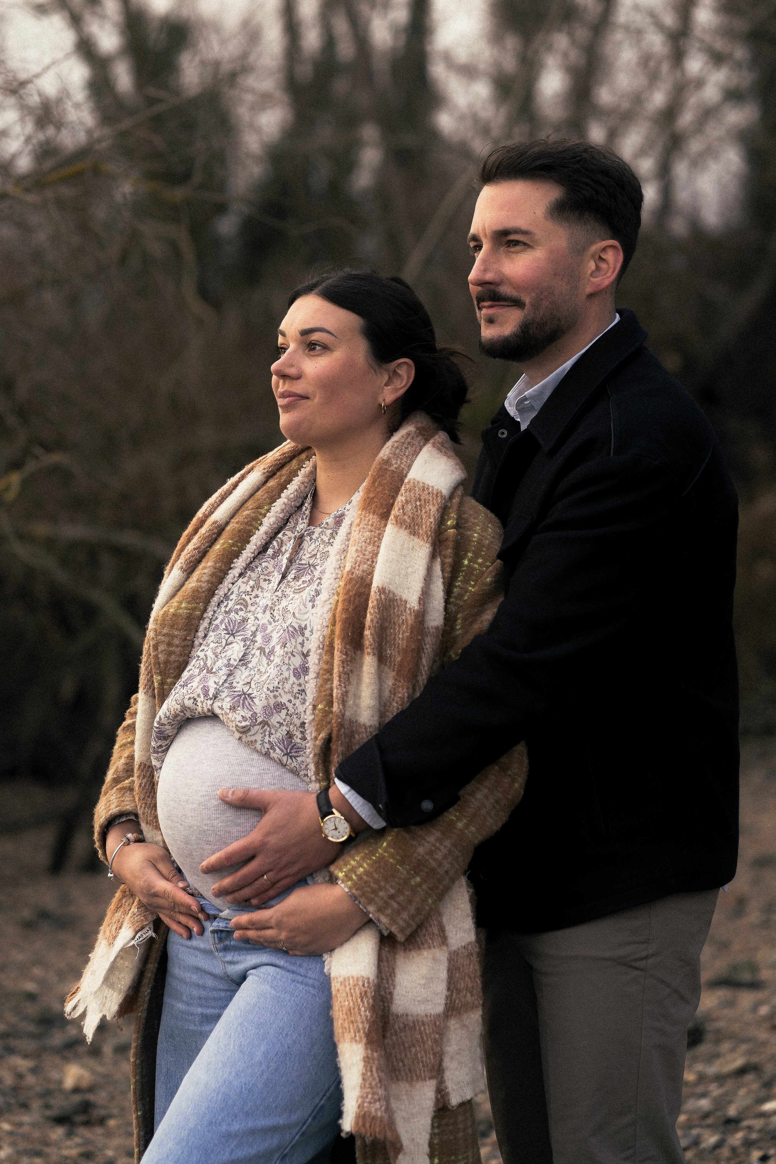 Un couple posant dans la nature, la femme est enceinte et porte un manteau à carreaux, l'homme se tient derrière elle, en la tenant par la taille et la montrant son ventre.