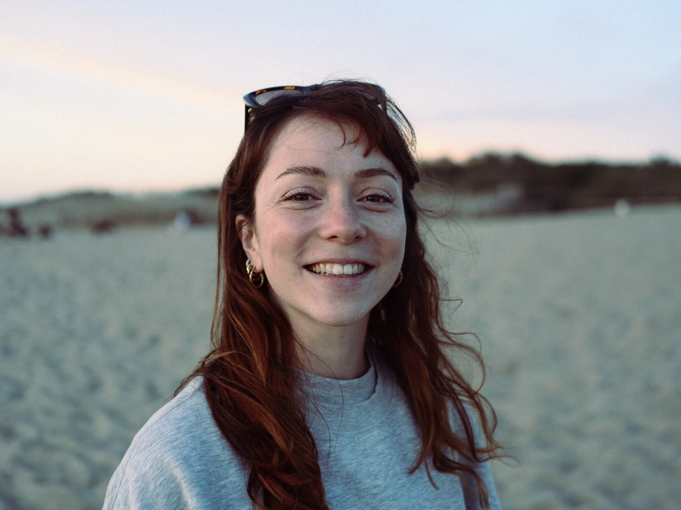 Une femme souriante avec des cheveux roux, portant des boucles d'oreilles dorées, se trouve à la plage lors du coucher du soleil sur la plage d'Anglet au Pays Basque.