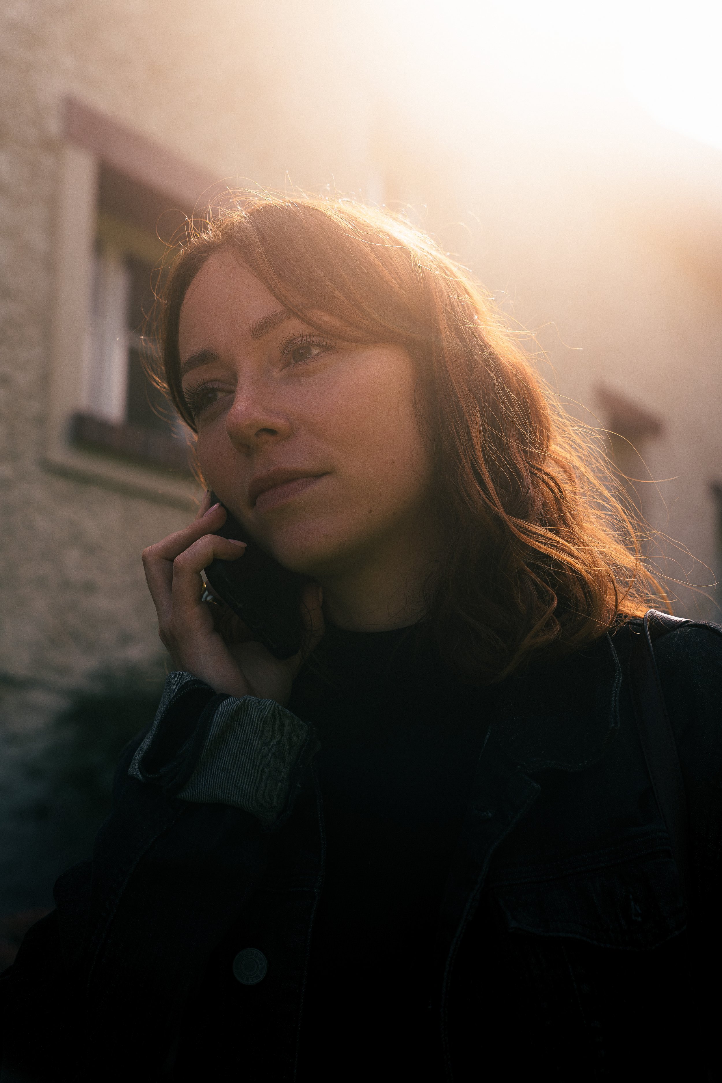 Jeune femme avec cheveux bruns, portant une veste en denim, parlant au téléphone devant une maison éclairée par la lumière du soleil couchant.