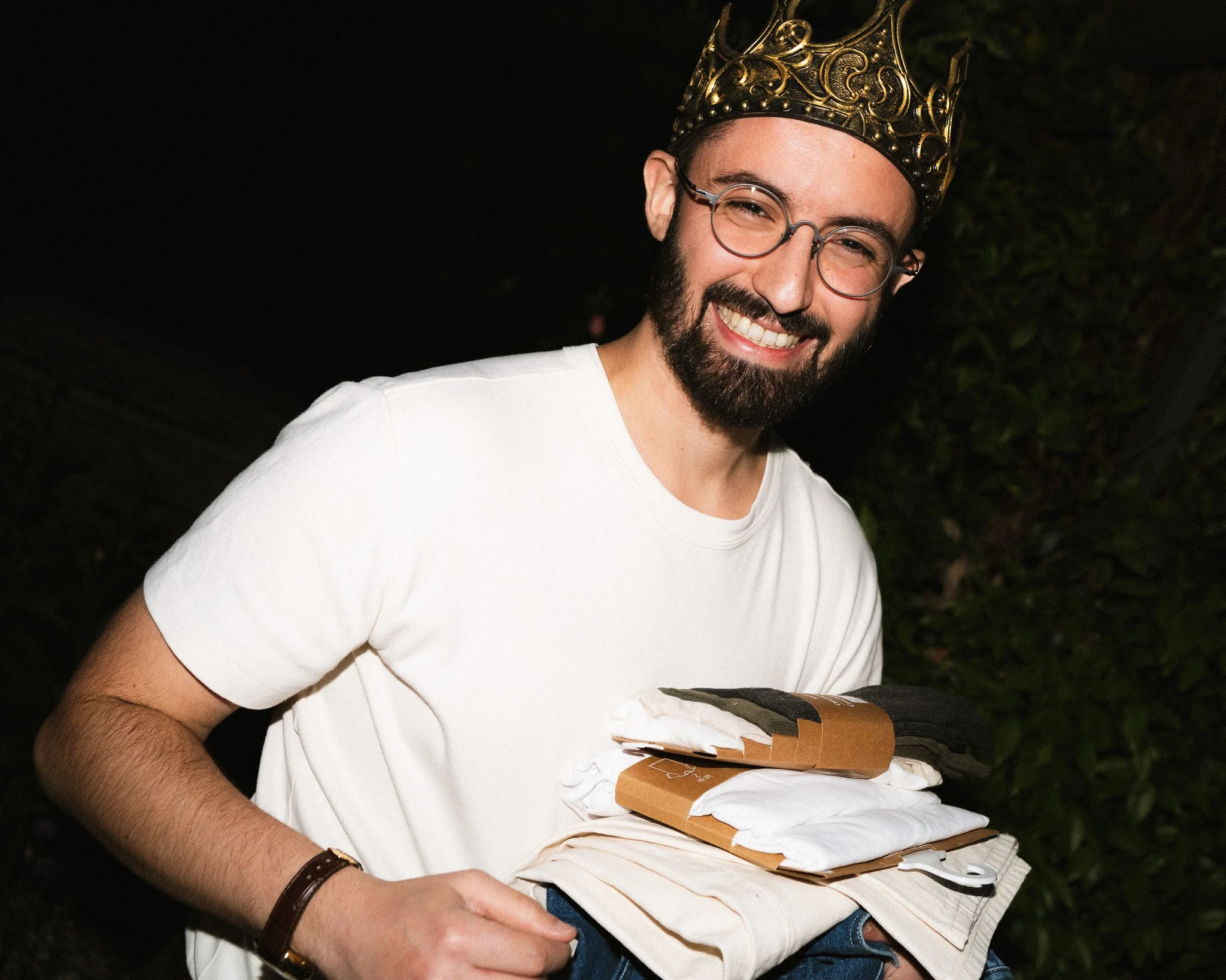 Un homme souriant portant une couronne dorée et lunettes, tenant des vêtements ou des papiers dans ses bras, à l'extérieur la nuit pour son anniversaire.