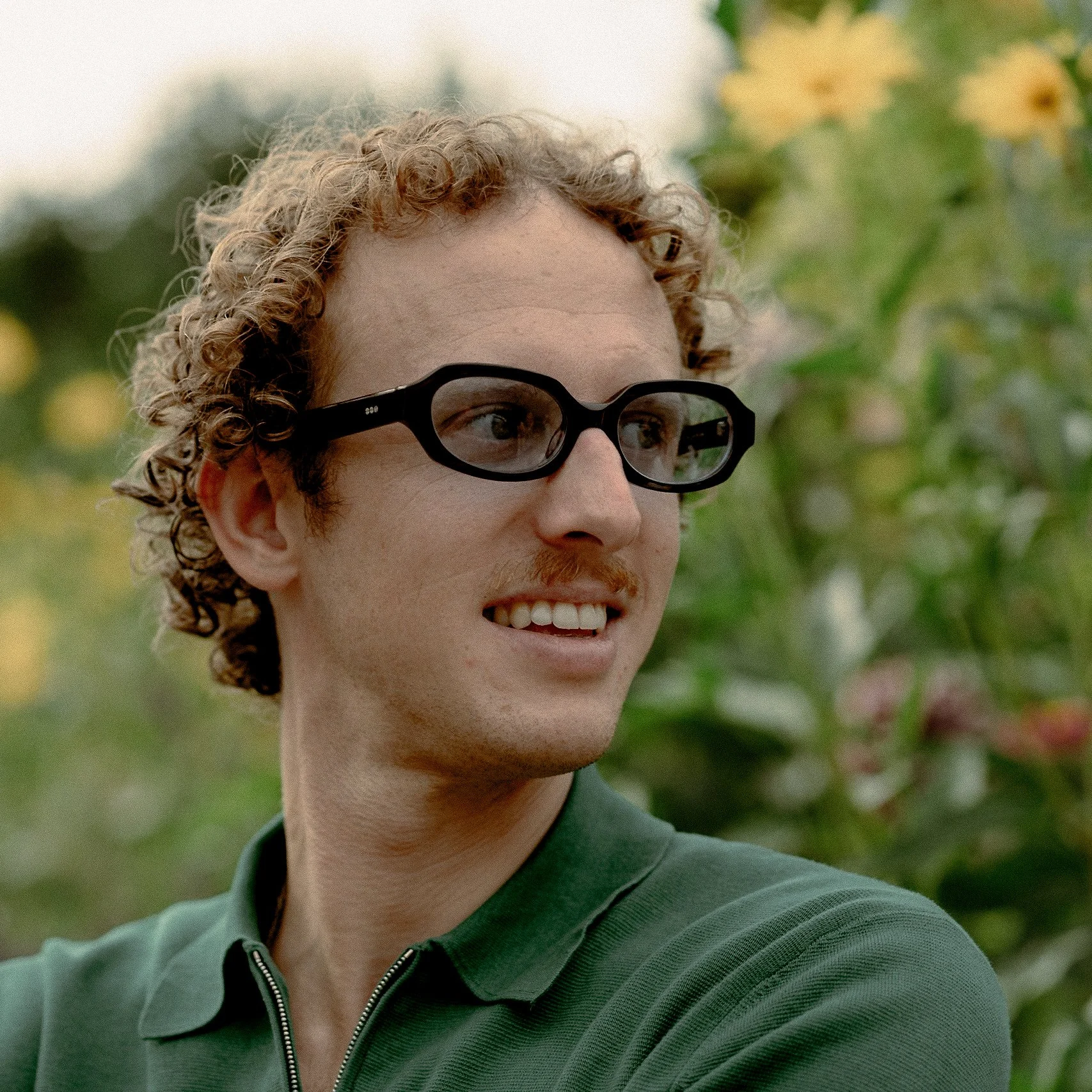 	
Un homme avec des cheveux bouclés de couleur claire porte des lunettes de protection et sourit, portant une veste verte. En arrière-plan, le jardin et la maison de Claude Monet à Giverny.