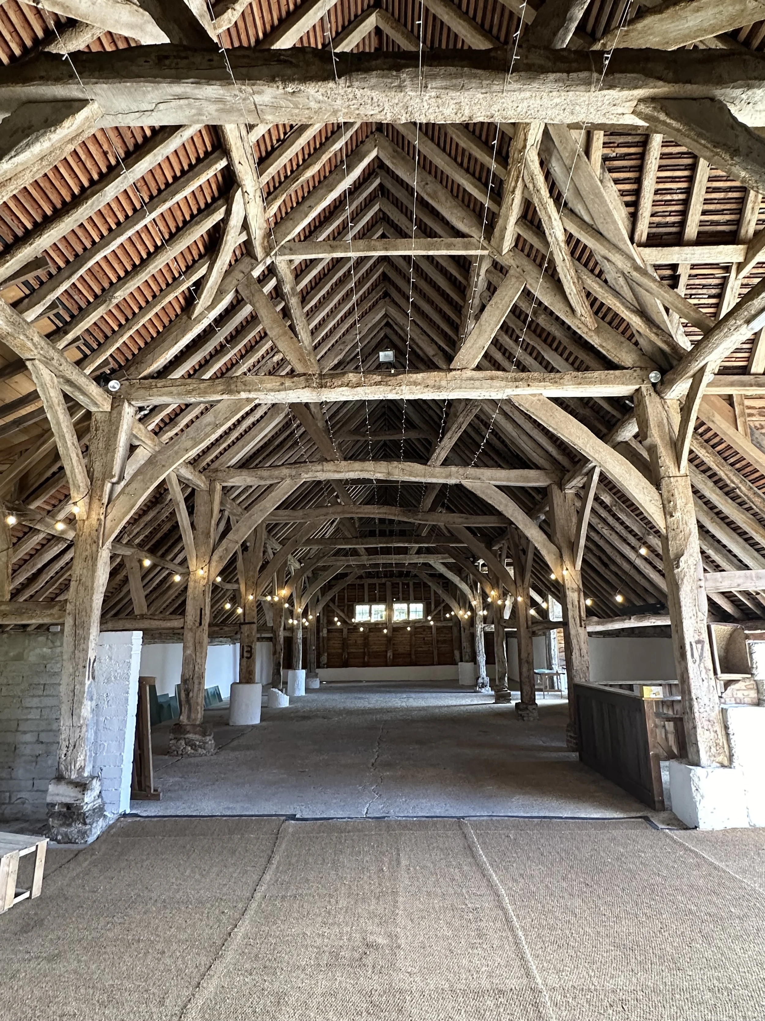 Interior of a rustic barn with exposed wooden beams, string lights, and a carpeted floor area.