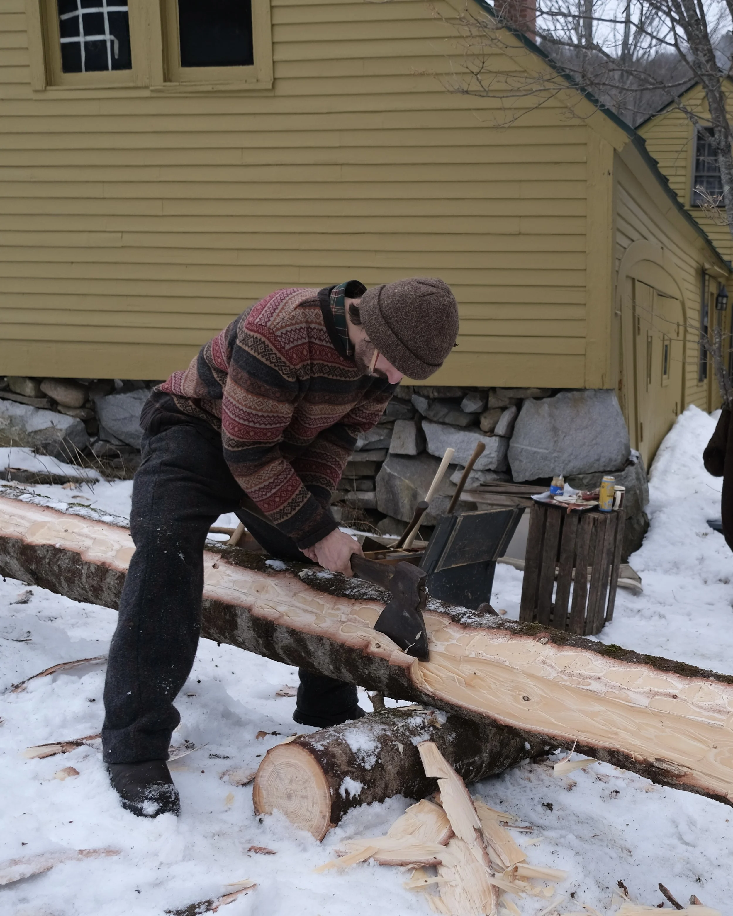 Nevan Carling hewing a large log with an axe outdoors in snow, with a yellow house in the background.
