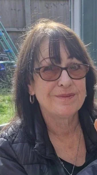 A woman with shoulder-length brown hair, wearing glasses and hoop earrings, is outdoors with a wooden fence and a blue structure in the background.