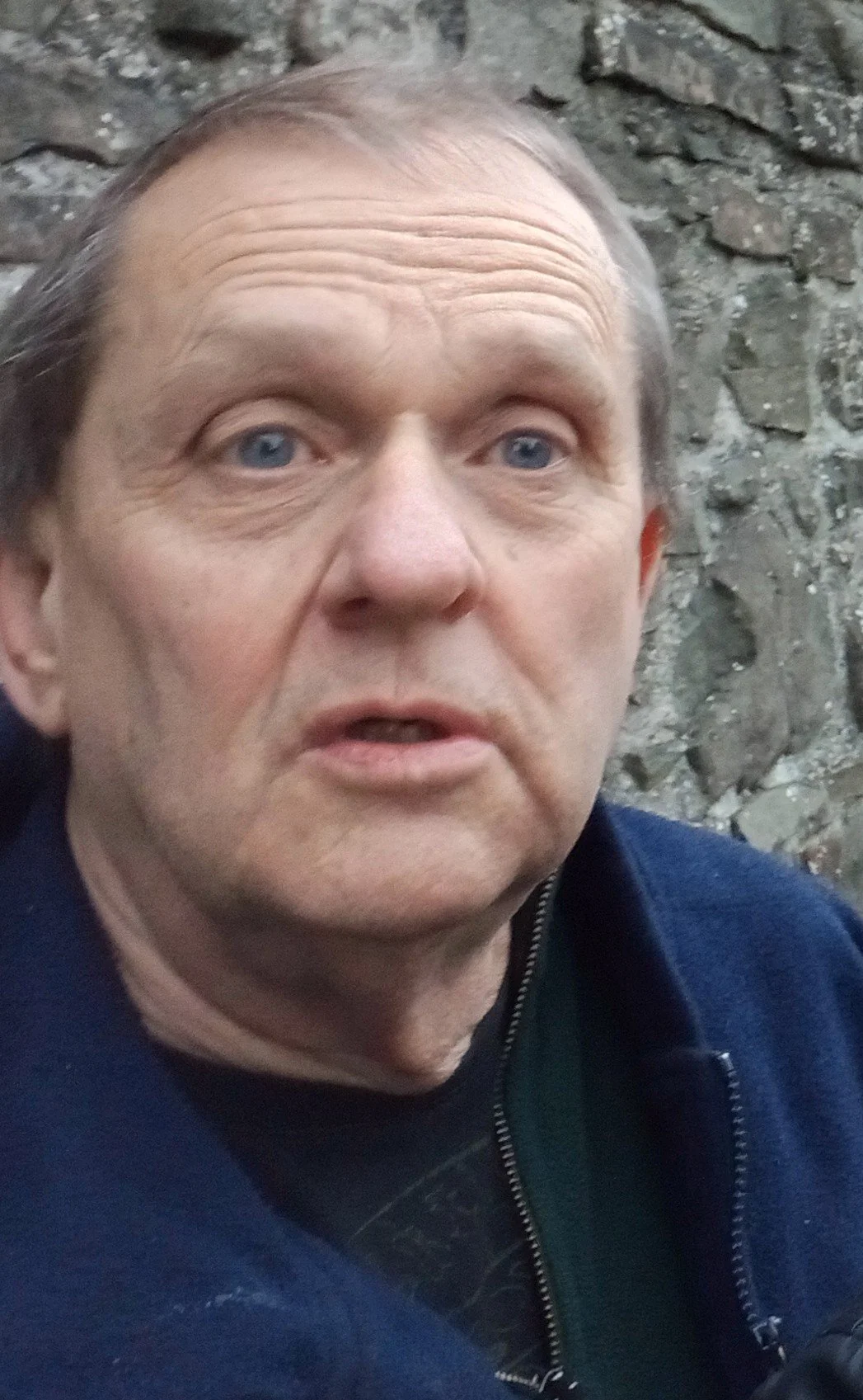 Close-up of a middle-aged man with light skin and blue eyes, wearing a dark jacket, standing outdoors in front of a stone wall.